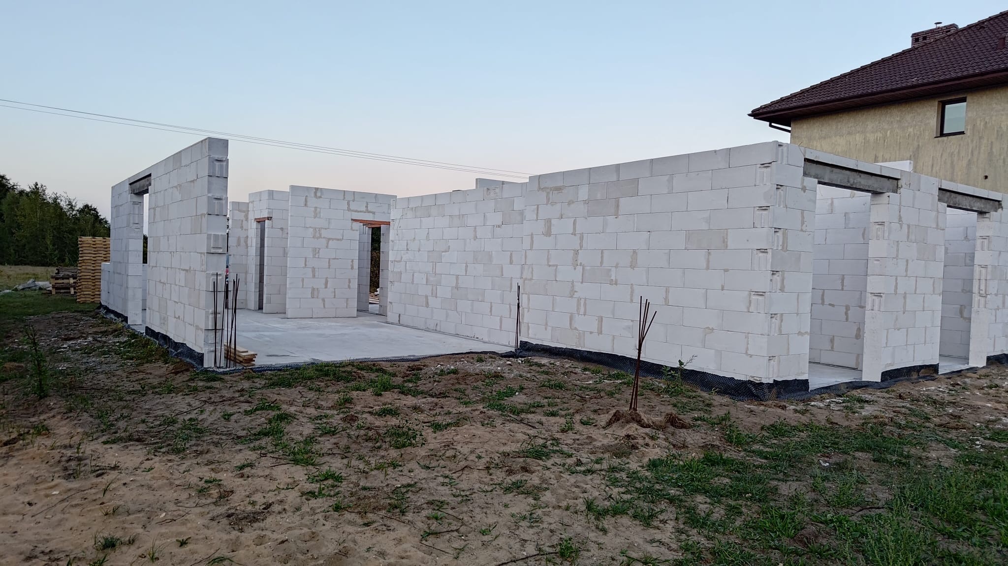 Wznoszone ściany z białych bloczków betonu komórkowego na fundamencie, widoczne pręty zbrojeniowe, w tle dom jednorodzinny i stos bloczków.
