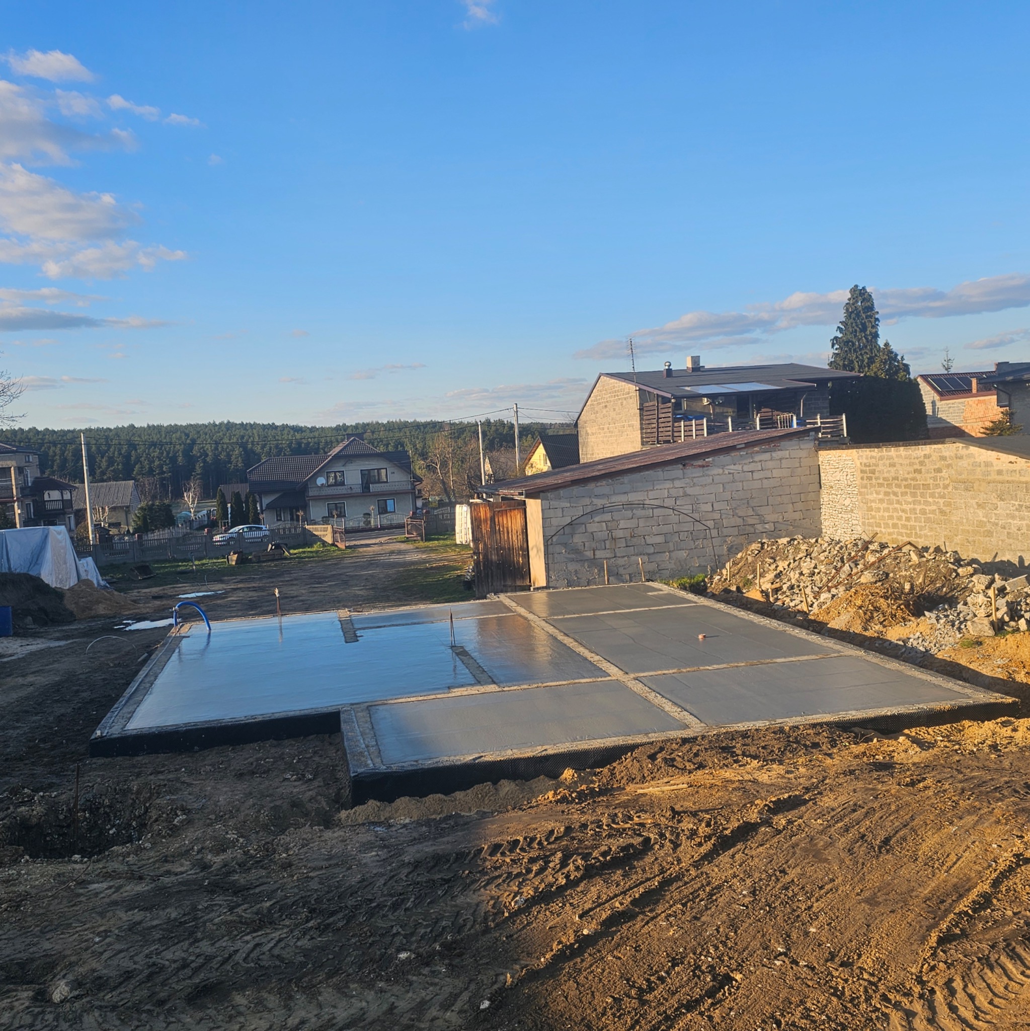Wylany betonowy fundament z widocznymi sekcjami, otoczony ziemią. W tle zabudowa jednorodzinna i błękitne niebo.