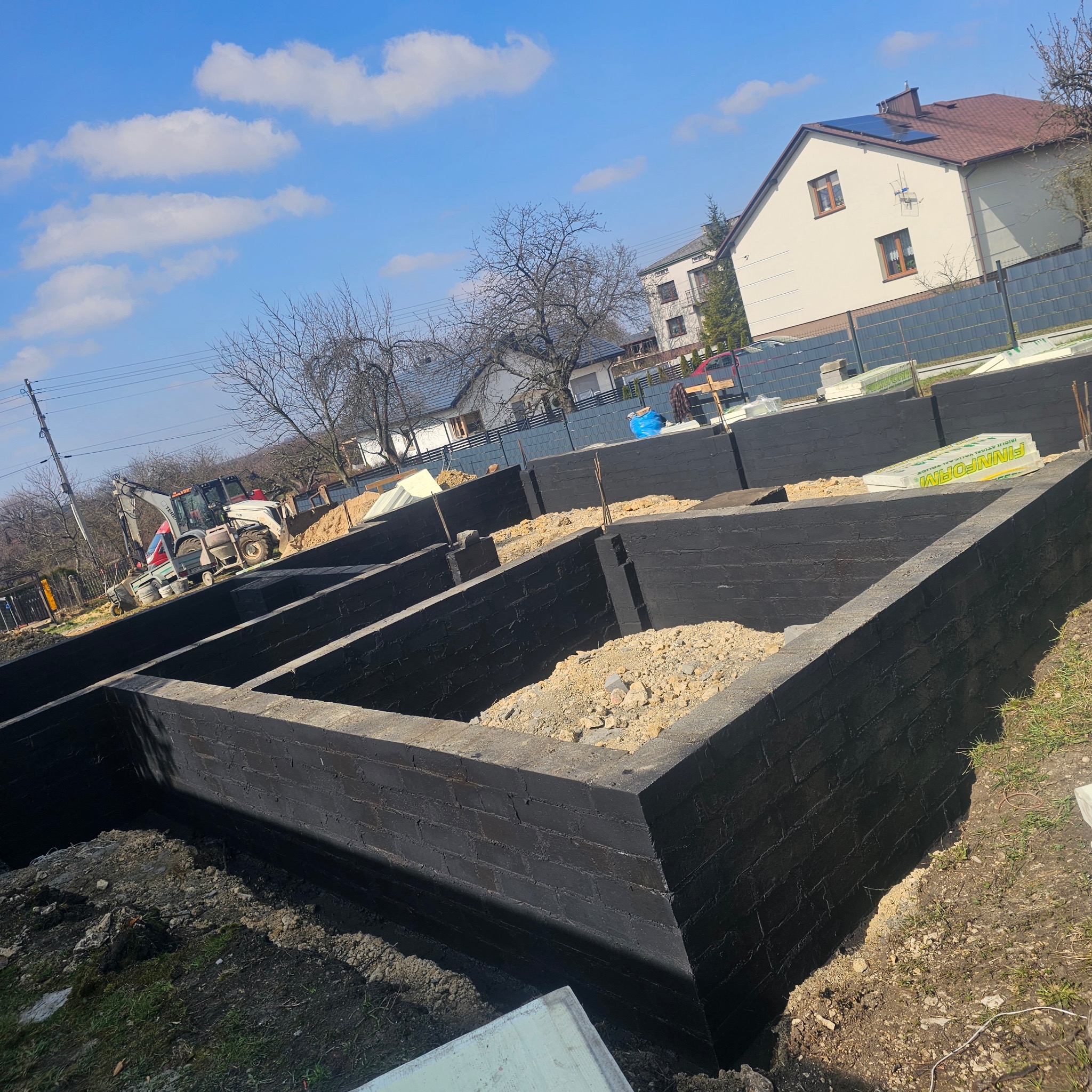 Zabezpieczone, czarną izolacją, murowane ściany fundamentowe, częściowo zasypane ziemią. W tle budynek mieszkalny i koparka. Błękitne niebo z obłokami.