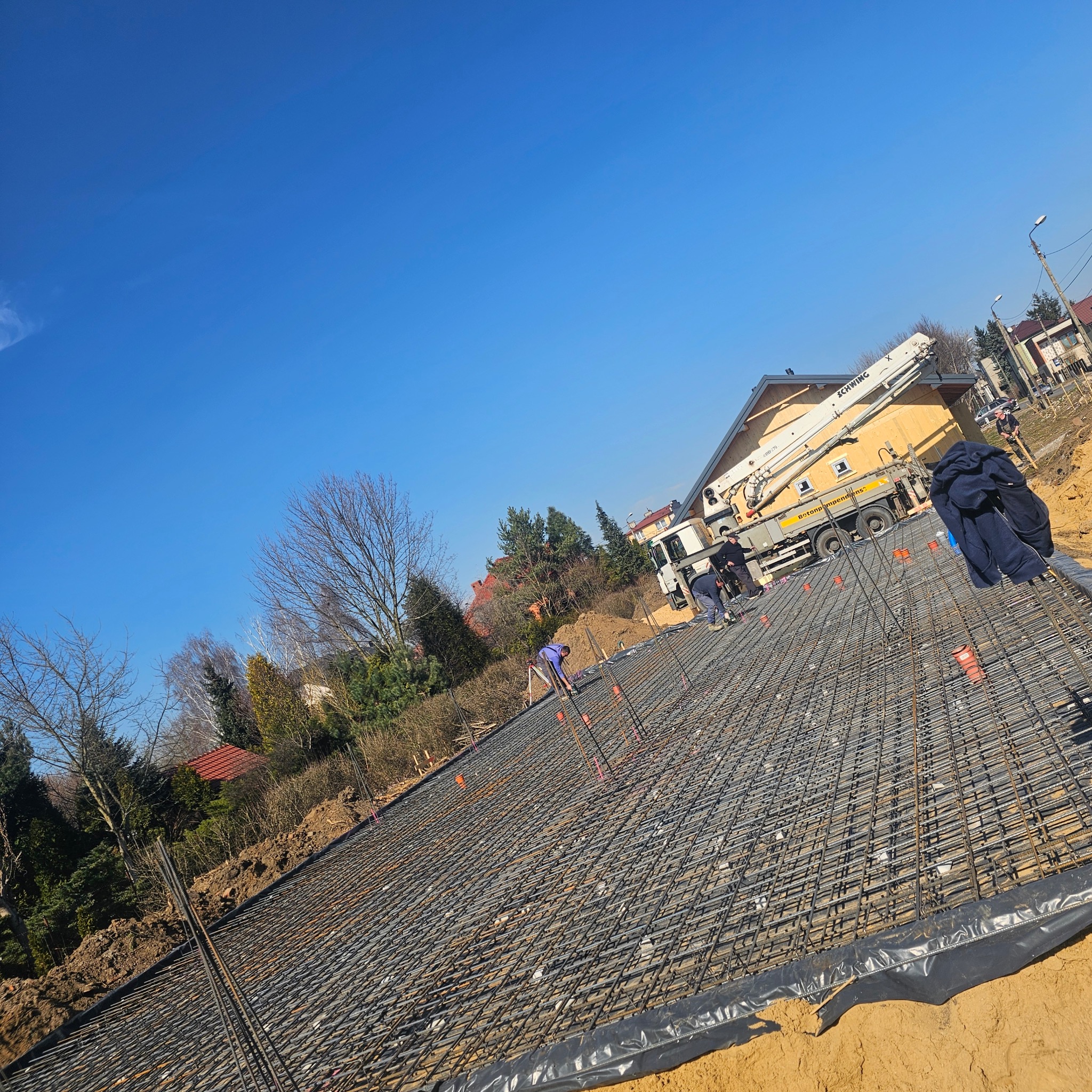 Zbrojenie fundamentów pod budynek jednorodzinny z użyciem pompy do betonu, widok z góry na siatkę zbrojeniową i pracowników. Słoneczny dzień.