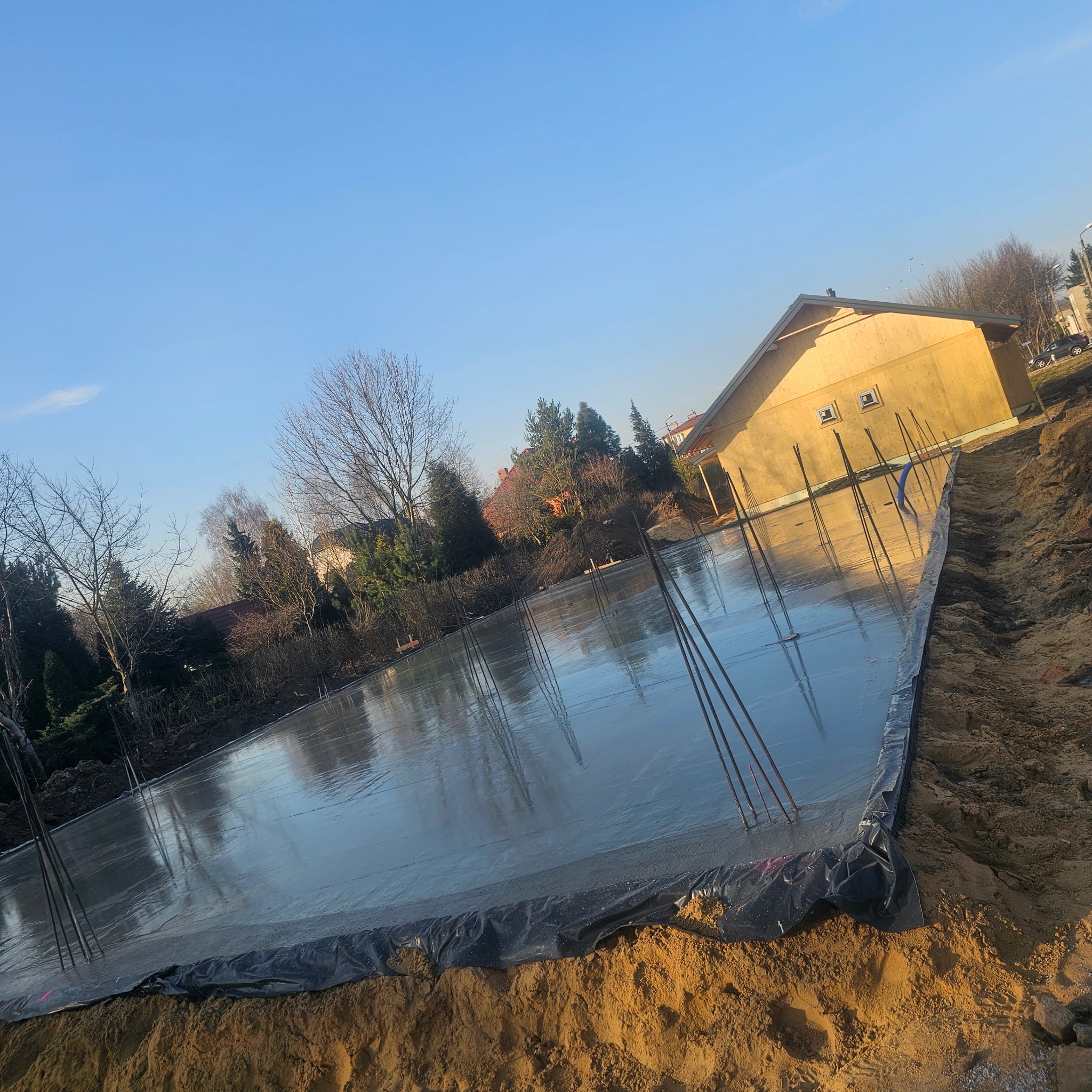 Świeżo wylany, gładki fundament pod budynek, z wystającymi prętami zbrojeniowymi, otoczony ziemią i folią. W tle widać budynek w stanie surowym.