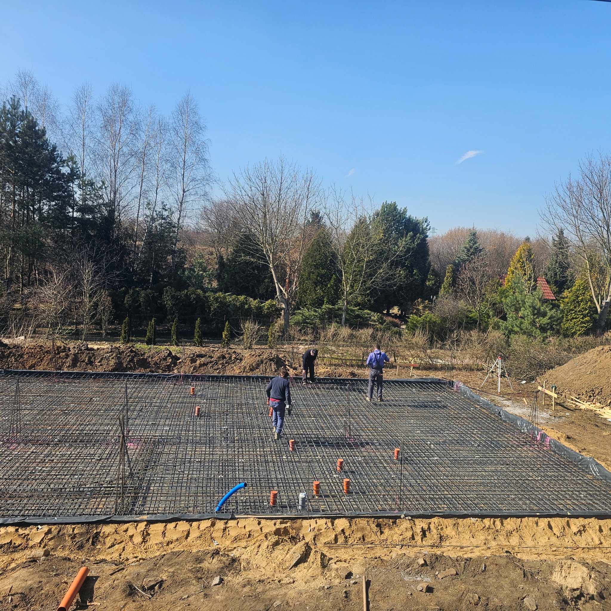 Zbrojenie płyty fundamentowej na przygotowanym terenie. Widoczne rury instalacyjne i pracownicy budowlani w trakcie pracy. Jasne niebo w tle.