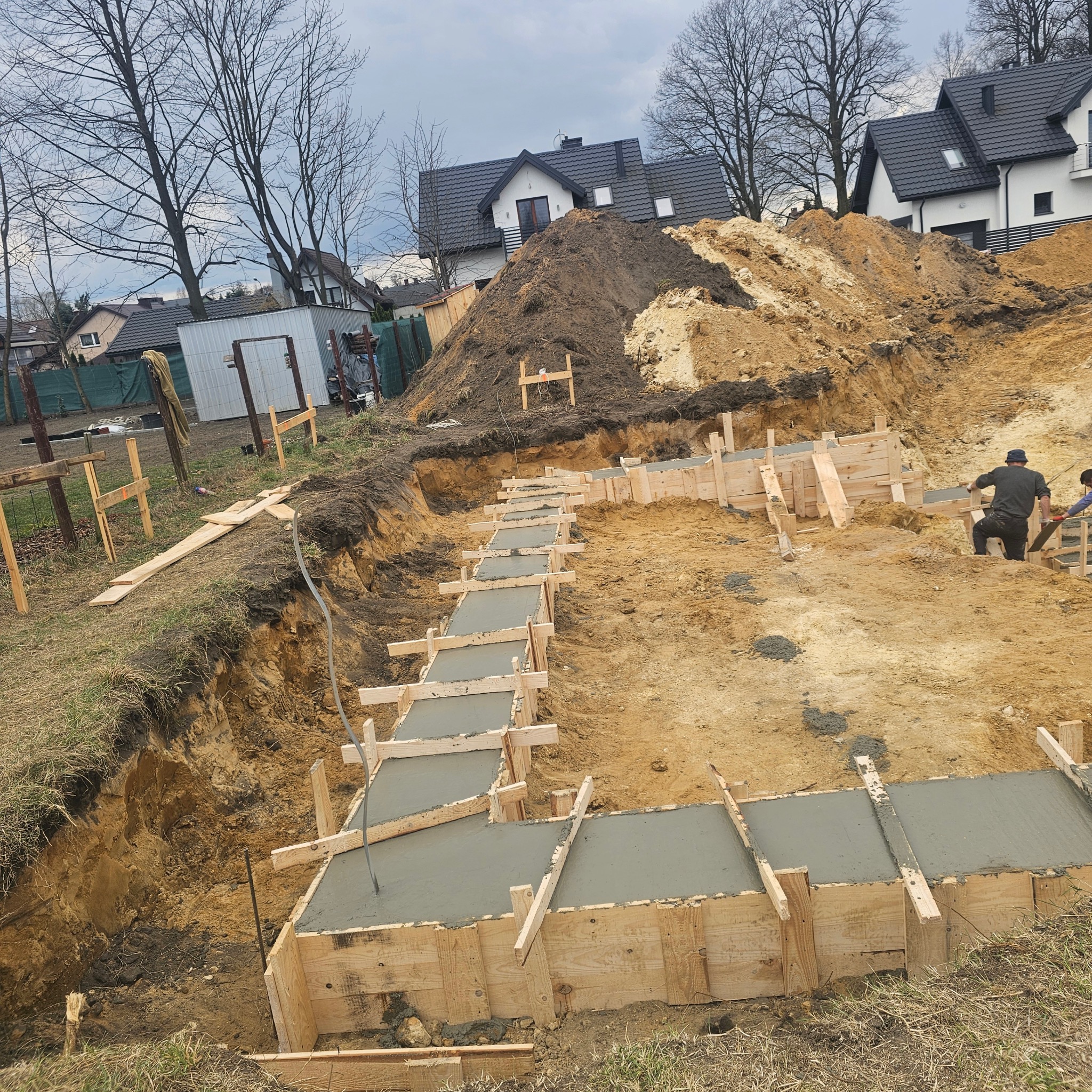 Zalewanie betonem szalunków fundamentów budynku jednorodzinnego. Widoczni pracownicy budowlani na tle wykopu i hałdy ziemi.