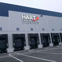 Zewnętrzny widok magazynu Hart Logistics z rampami załadunkowymi oznaczonymi numerami 15-21 i zaparkowaną naczepą ciężarówki przy rampie 21.
