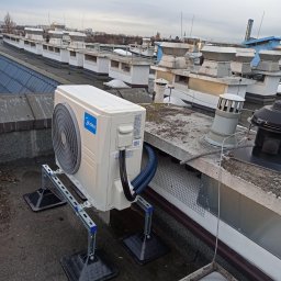 Trodos sp. z o. o - Biała jednostka zewnętrzna klimatyzacji Midea na metalowej konstrukcji wsporczej, zainstalowana na płaskim dachu budynku z licznymi wywietrznikami, widok z boku.