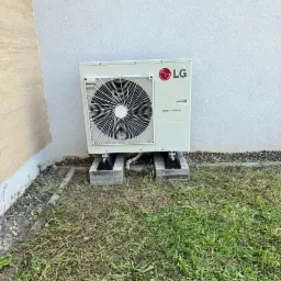 Zewnętrzna jednostka klimatyzacji LG Smart Inverter, zamontowana na betonowych bloczkach przy ścianie budynku, otoczona trawnikiem.