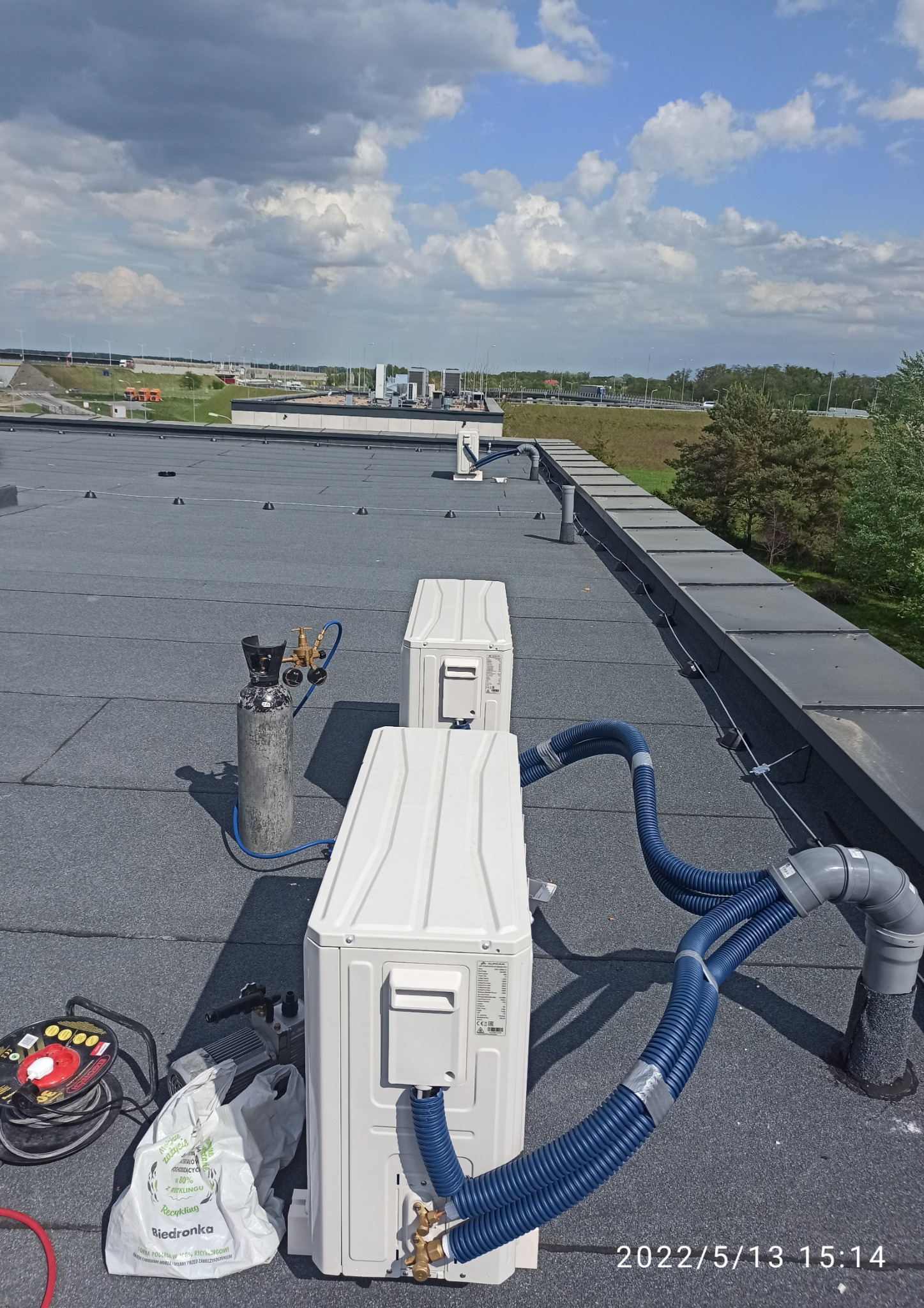 Dwa białe klimatyzatory na płaskim, bitumicznym dachu budynku, połączone niebieskimi przewodami; obok butla z gazem i narzędzia, w tle widok na autostradę i niebo z chmurami.