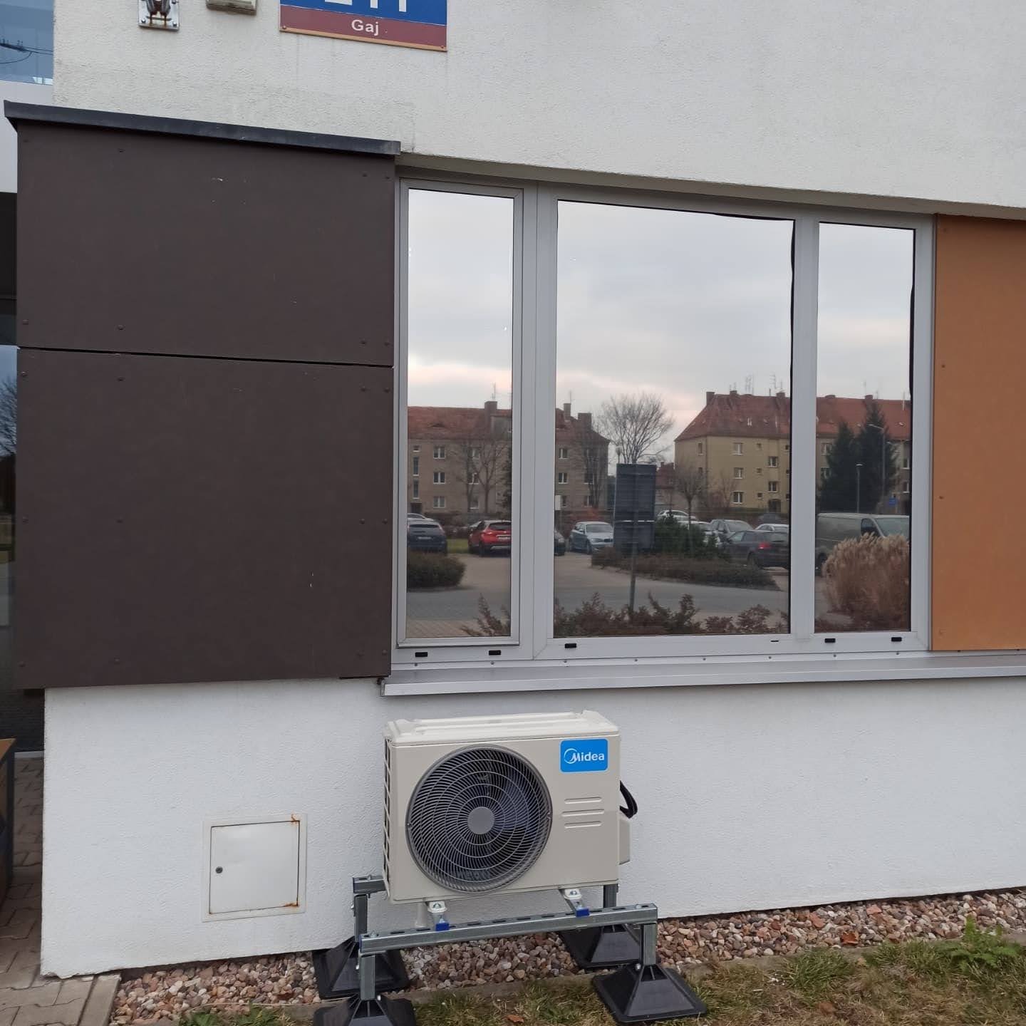 Zewnętrzna jednostka klimatyzacji Midea zamontowana na metalowej konstrukcji przy ścianie budynku z widocznym oknem odbijającym otoczenie.