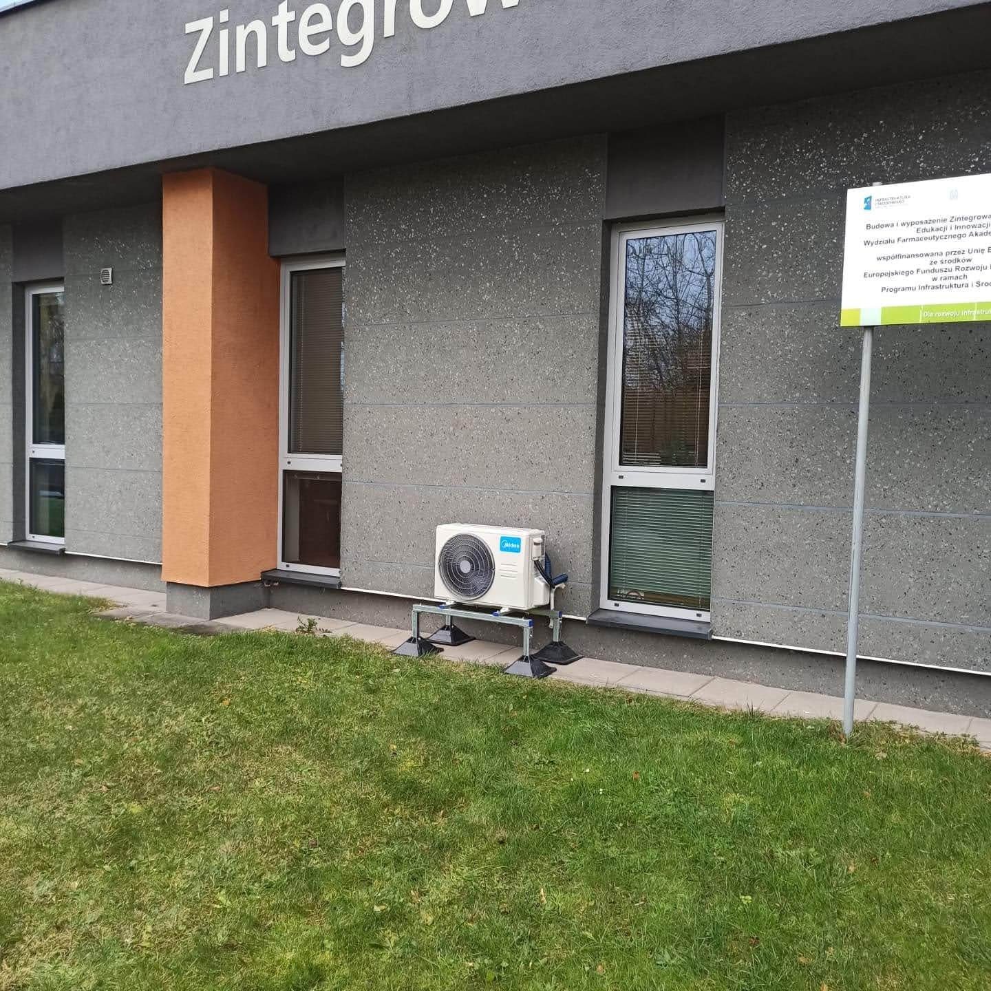 Zewnętrzna jednostka klimatyzacji Midea zamontowana na metalowej konstrukcji przed budynkiem z szarą elewacją i oknami, widoczny trawnik i tablica informacyjna.