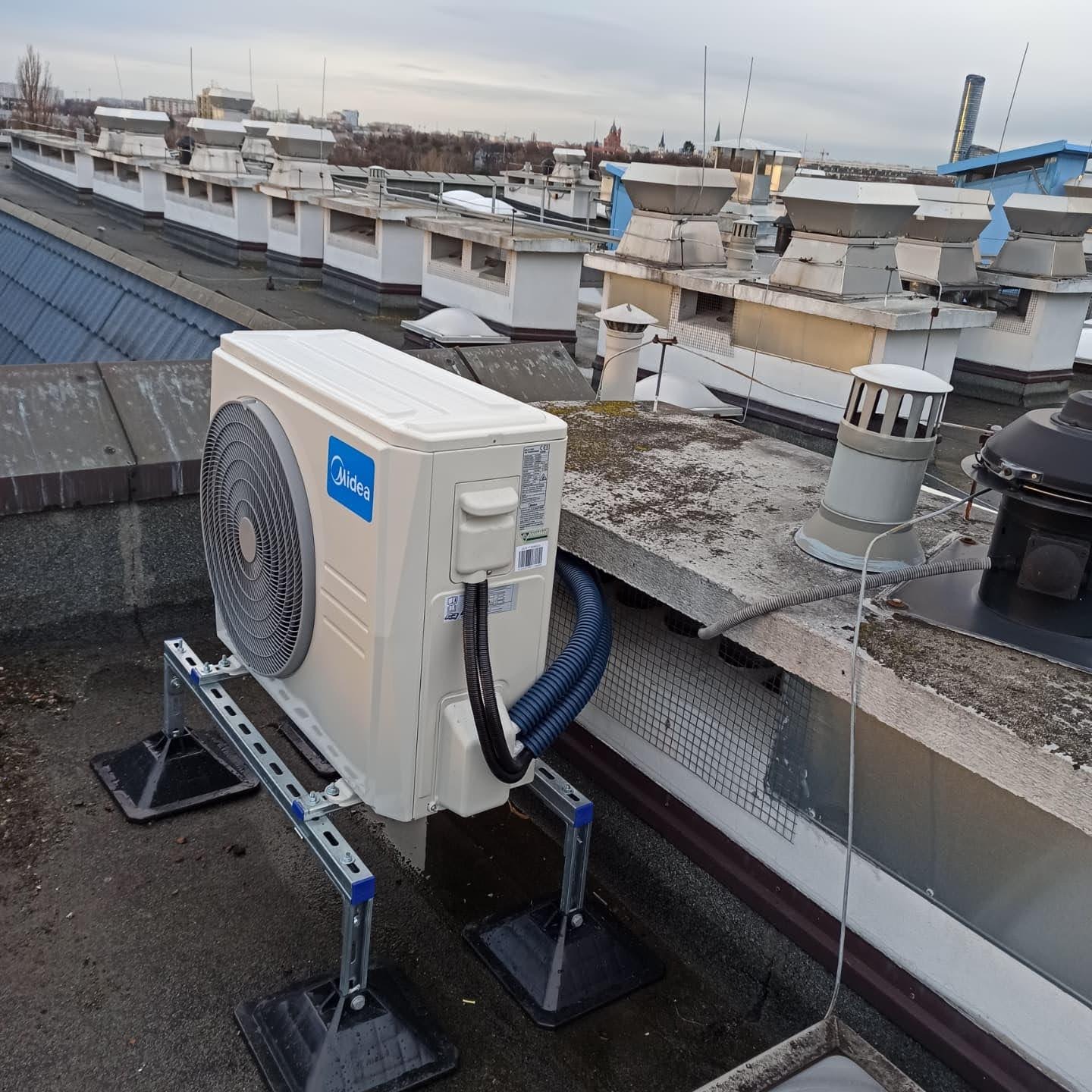 Biała jednostka zewnętrzna klimatyzacji Midea na metalowej konstrukcji wsporczej, zainstalowana na płaskim dachu budynku z licznymi wywietrznikami, widok z boku.