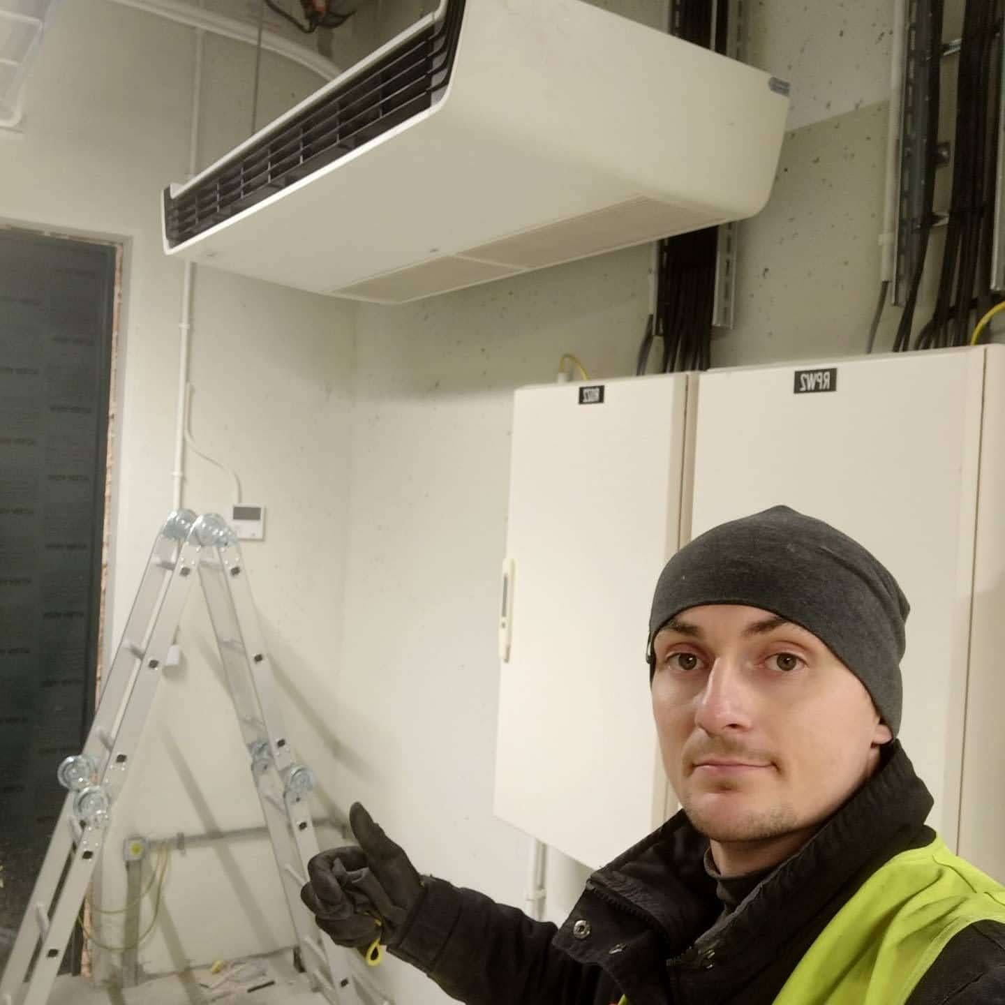Mężczyzna w kamizelce odblaskowej i czapce, z kciukiem w górę, na tle zamontowanej klimatyzacji kanałowej w pomieszczeniu biurowym, z widoczną drabiną i szafami elektrycznymi.