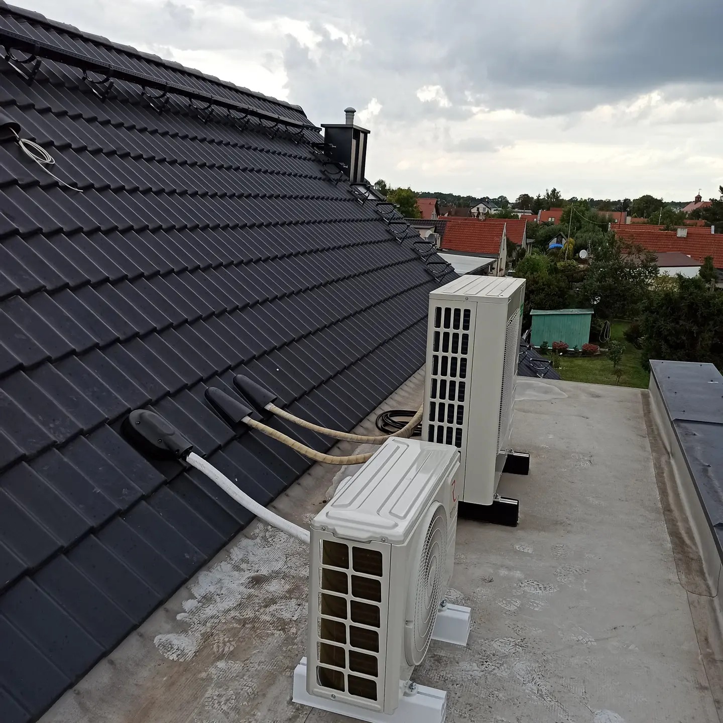 Dwa klimatyzatory zewnętrzne zamontowane na płaskim dachu budynku mieszkalnego z widocznymi przewodami biegnącymi do dachu pokrytego czarną dachówką, w tle zabudowa jednorodzinna i pochmurne niebo.