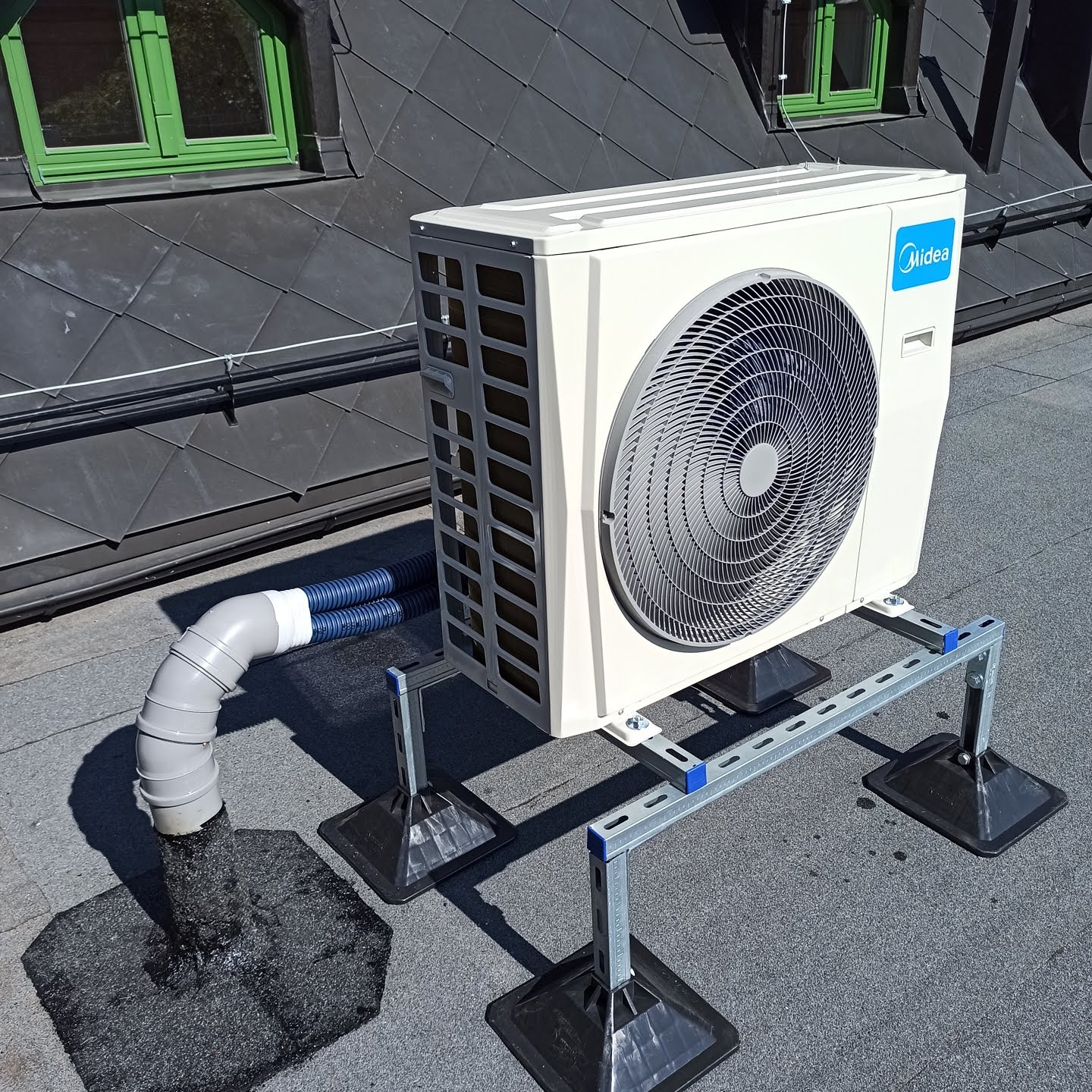 Zewnętrzna jednostka klimatyzacji Midea zamontowana na dachu z szarej dachówki, z widocznymi przewodami odprowadzającymi skropliny i ramą montażową.