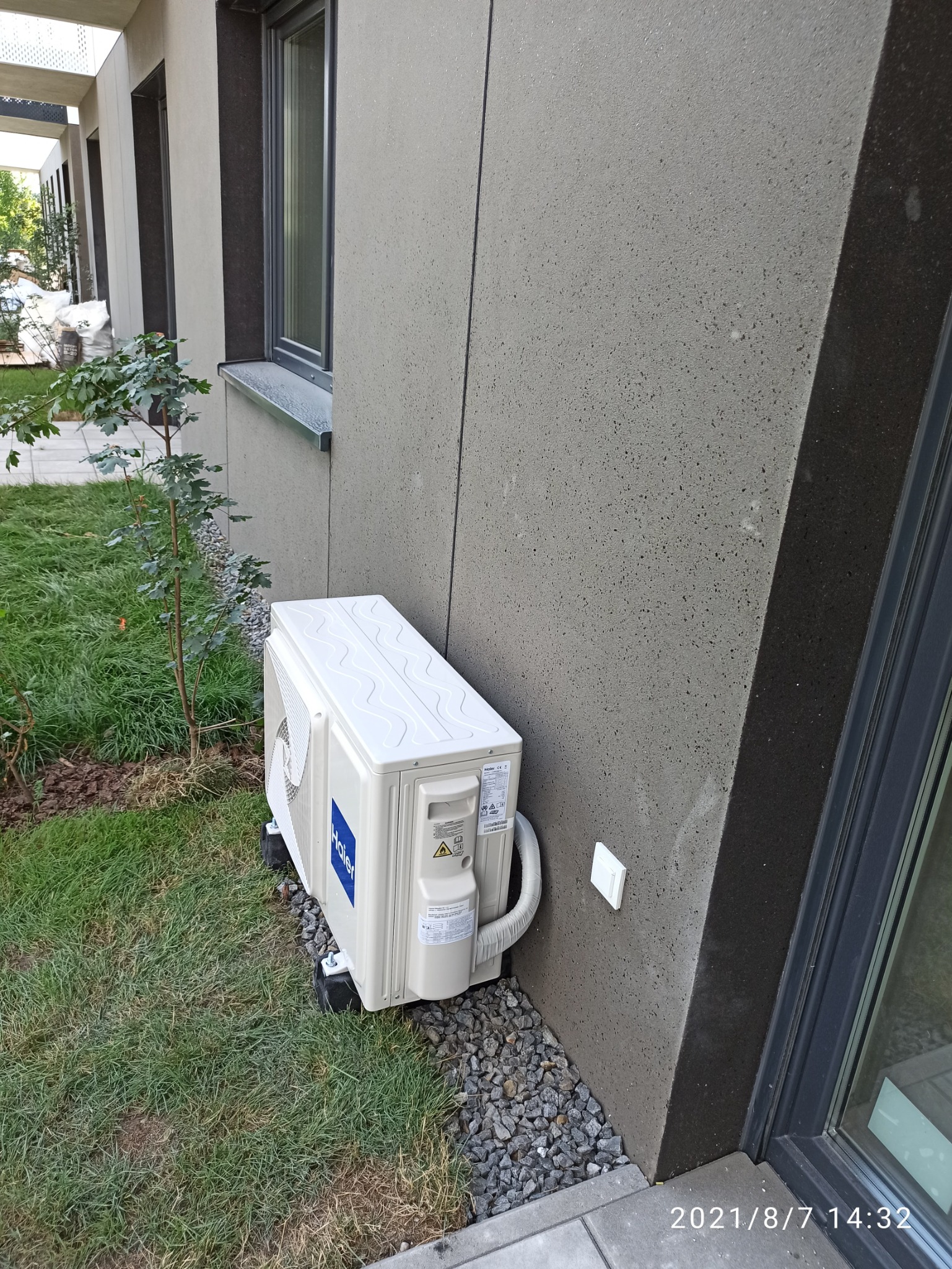 Zewnętrzna jednostka klimatyzacji Haier zamontowana na trawniku przy ścianie budynku z szarą elewacją, widoczny fragment okna i młode drzewko.