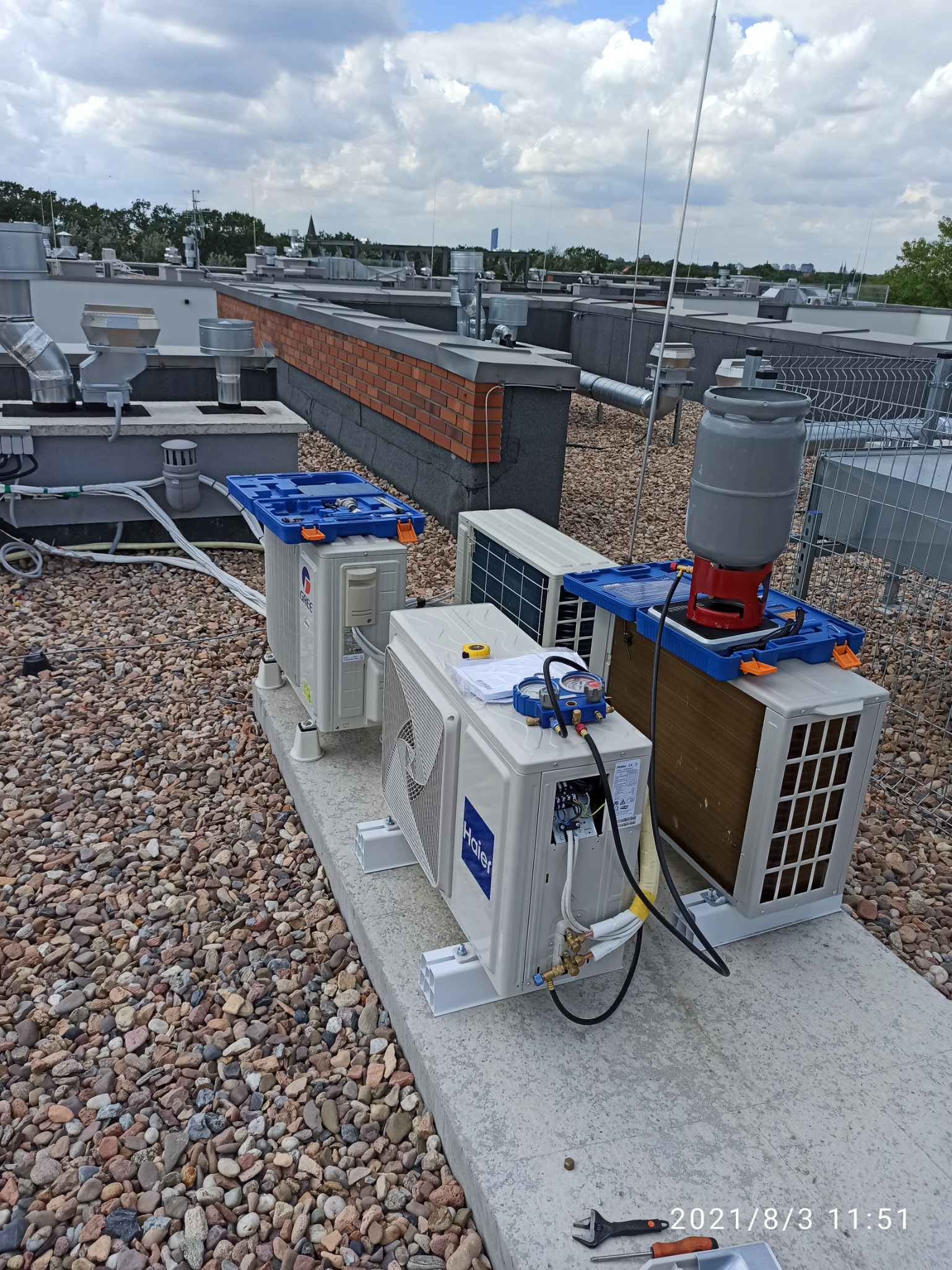 Instalacja klimatyzatorów Haier i Gree na dachu budynku, widoczne podłączenia, narzędzia i butla z gazem chłodniczym.