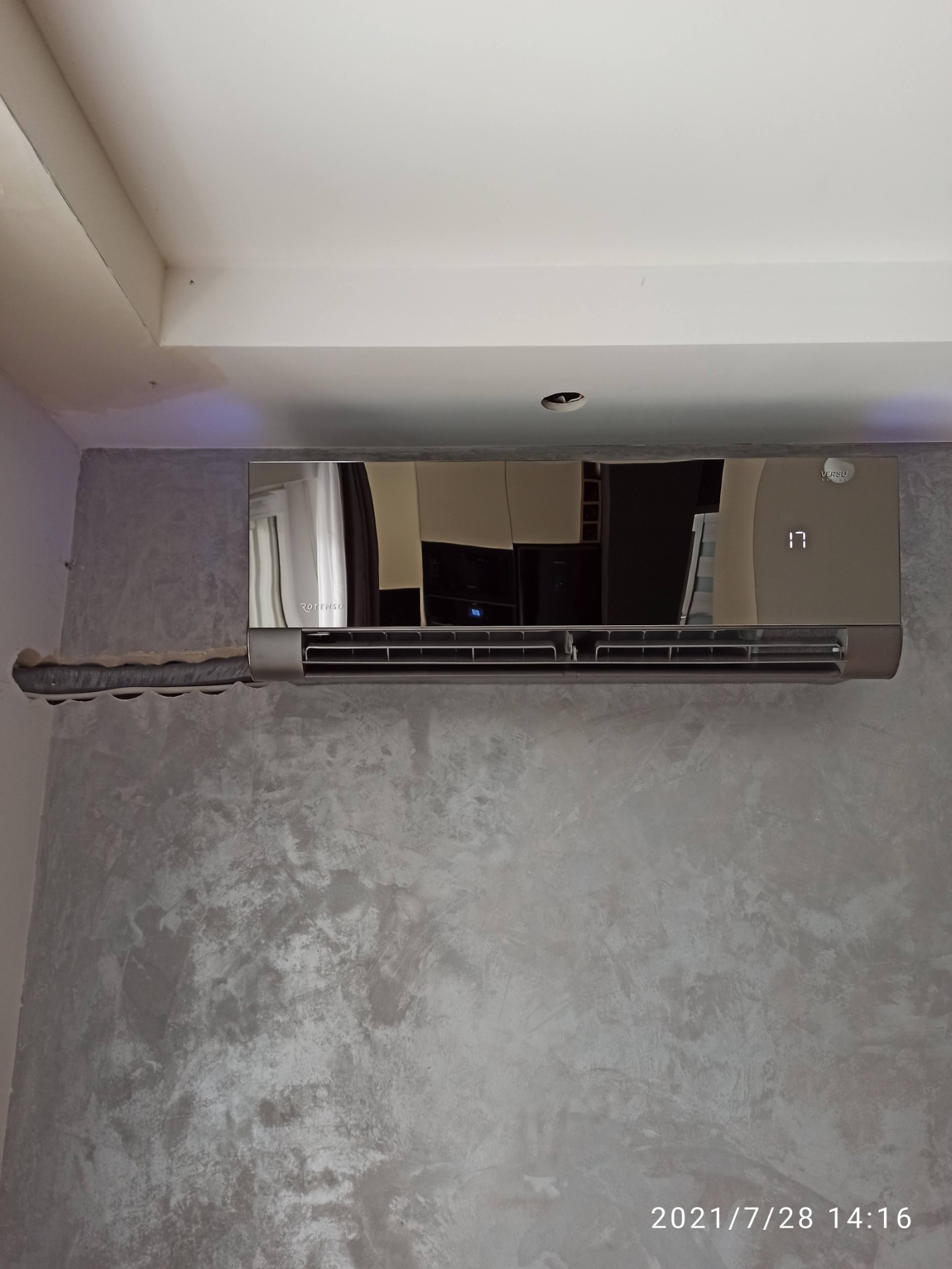 Szary klimatyzator z wyświetlaczem temperatury zamontowany na ścianie o fakturze betonu, zabezpieczony przed kurzem podczas remontu.