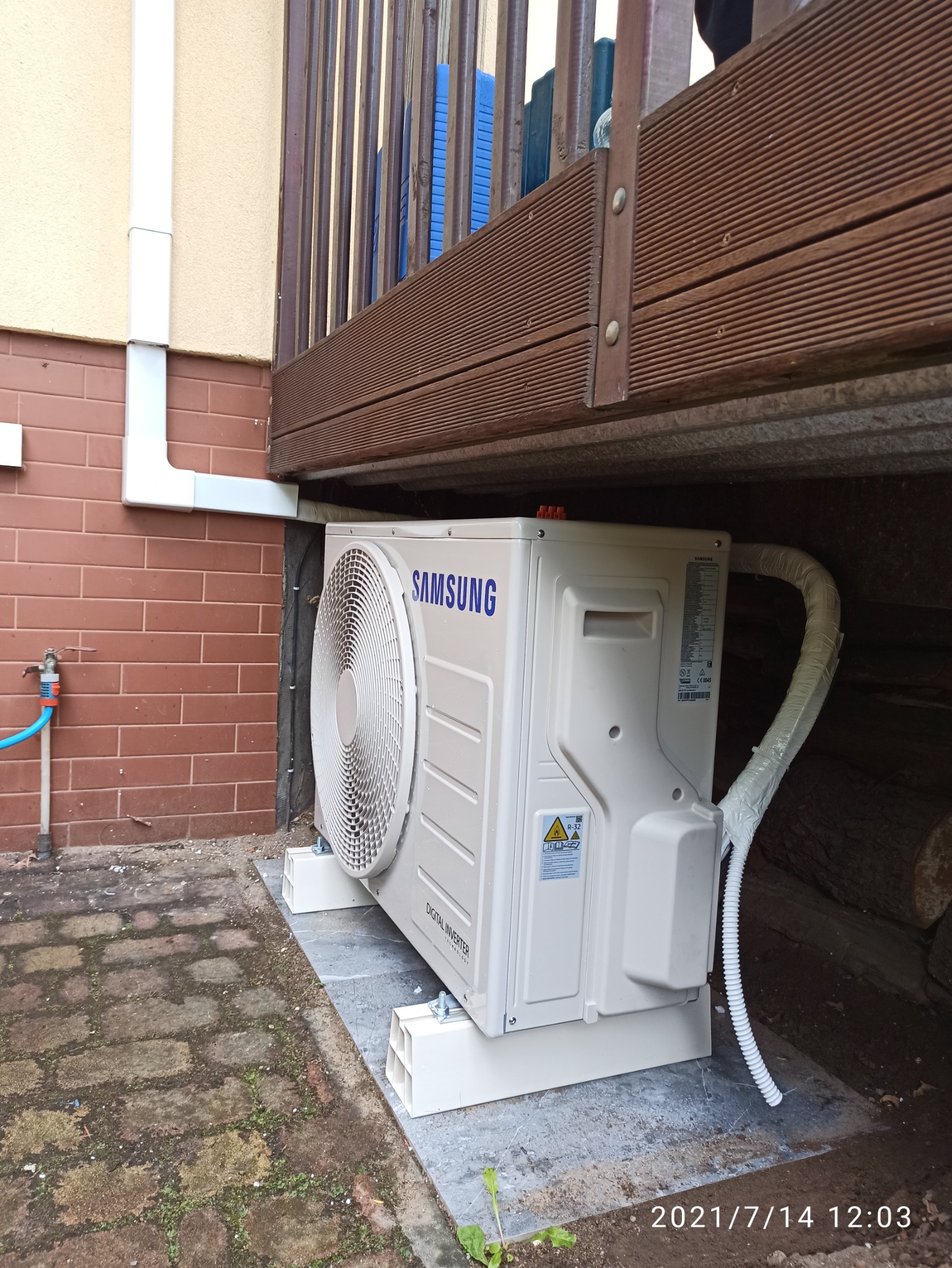 Zewnętrzna jednostka klimatyzacji Samsung Digital Inverter zamontowana na podeście z pustaków, z widocznymi rurami w białej izolacji, na tle ceglanej ściany i drewnianego tarasu.