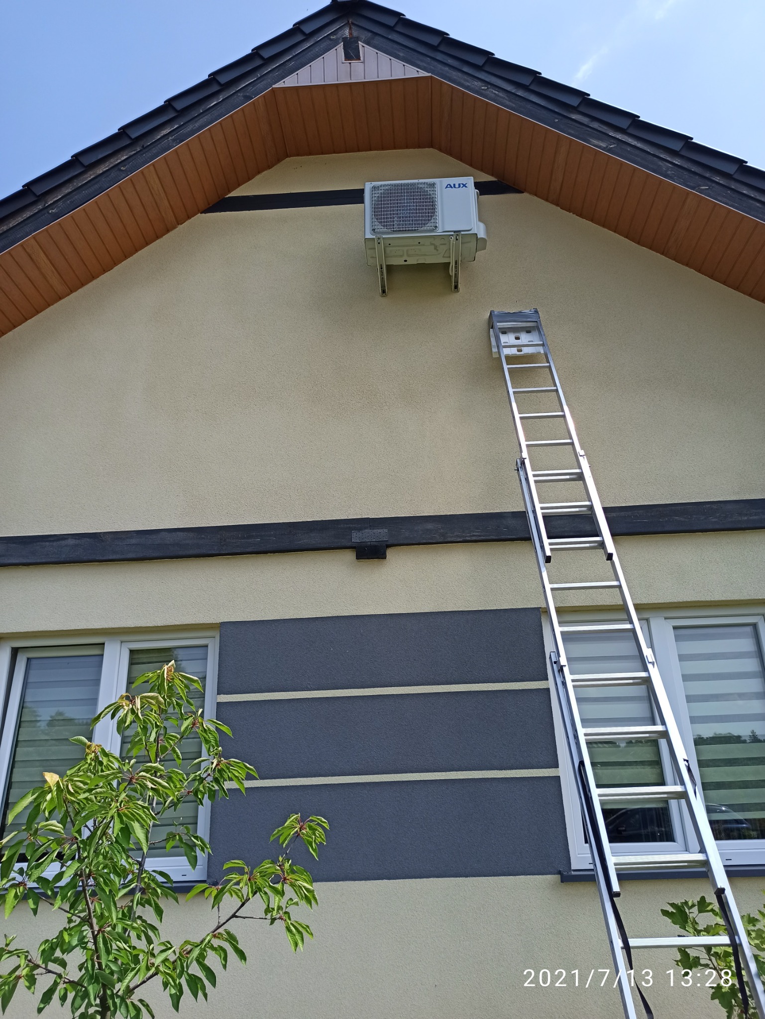 Biała jednostka zewnętrzna klimatyzacji AUX zamontowana na elewacji domu, obok przystawiona aluminiowa drabina.