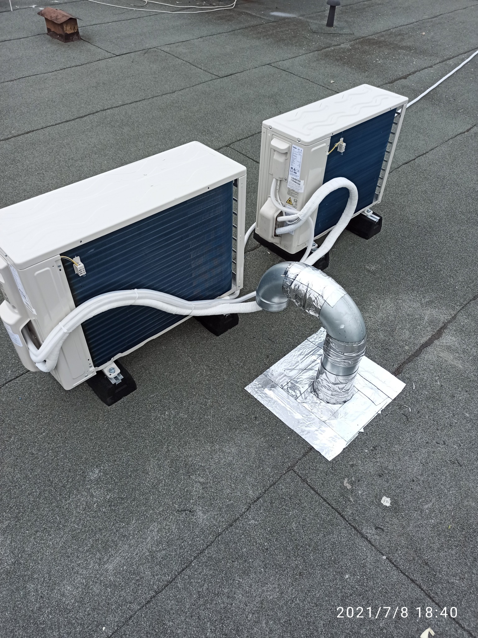 Dwa agregaty klimatyzacyjne na dachu pokrytym papą, jeden z podłączonym srebrnym przewodem wentylacyjnym zabezpieczonym folią aluminiową.