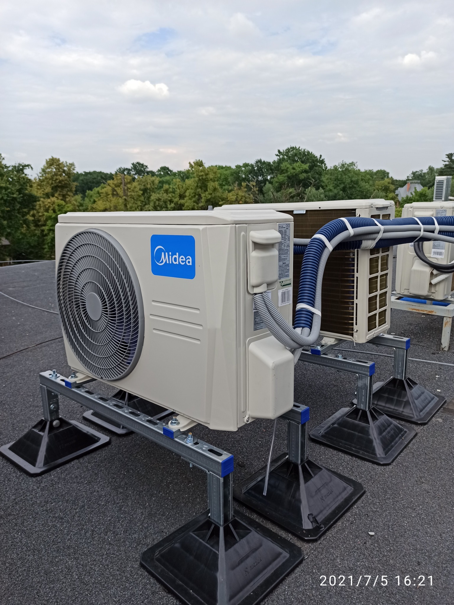 Zewnętrzne jednostki klimatyzacji Midea zamontowane na dachu budynku, widoczne rury i przewody, w tle drzewa i pochmurne niebo.