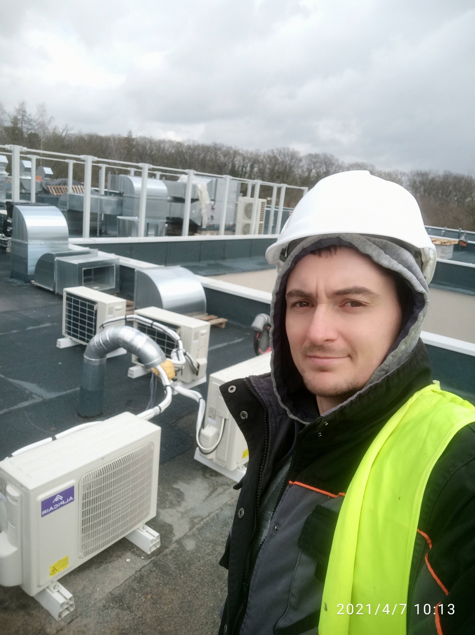 Instalator w kasku i kamizelce odblaskowej robi selfie na dachu budynku z zamontowanymi jednostkami zewnętrznymi klimatyzacji i kanałami wentylacyjnymi.