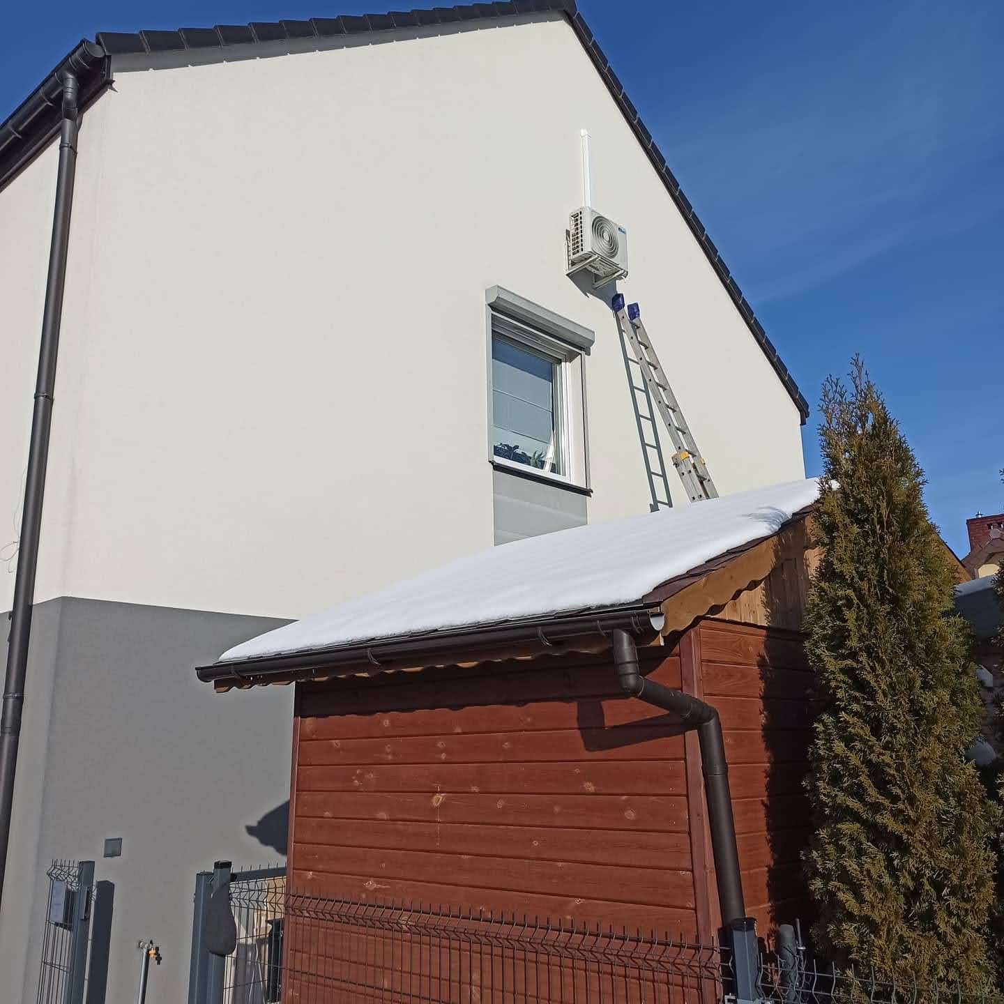 Biała jednostka zewnętrzna klimatyzacji zamontowana na ścianie domu, wsparta drabiną aluminiową, z widocznym odpływem pionowym, na tle błękitnego nieba i fragmentu drewnianej szopy ze śniegiem...
