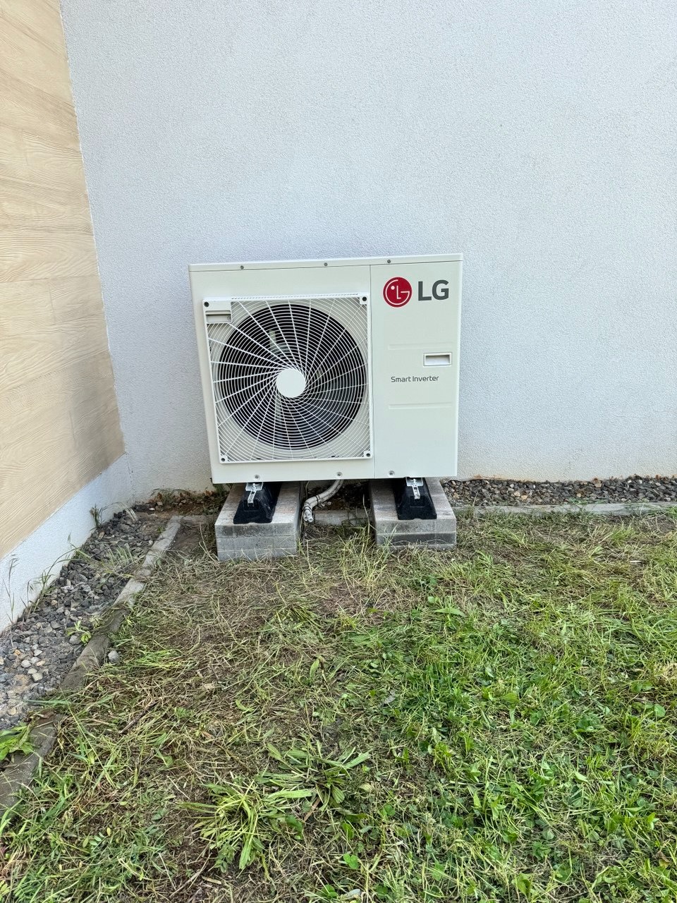 Zewnętrzna jednostka klimatyzacji LG Smart Inverter, zamontowana na betonowych bloczkach przy ścianie budynku, otoczona trawnikiem.