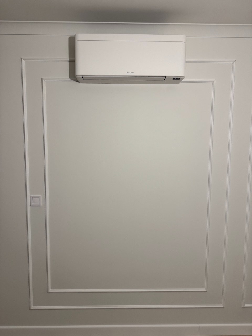 Biała klimatyzacja Daikin zamontowana na ścianie z dekoracyjnymi listwami w stylu klasycznym. Widoczny włącznik światła po lewej stronie.