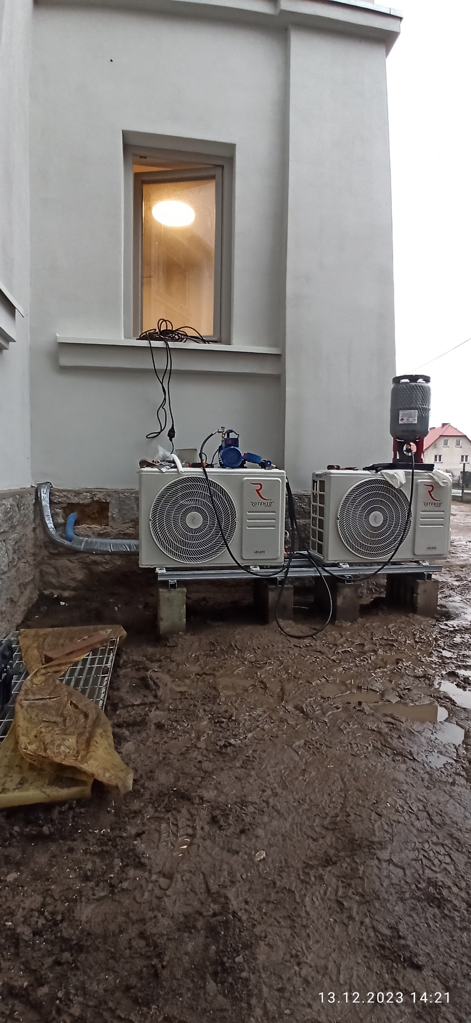 Dwa zewnętrzne moduły klimatyzacji Rotenso ustawione na betonowych blokach przed budynkiem, z widocznymi przewodami i narzędziami do instalacji na tle błotnistego terenu.