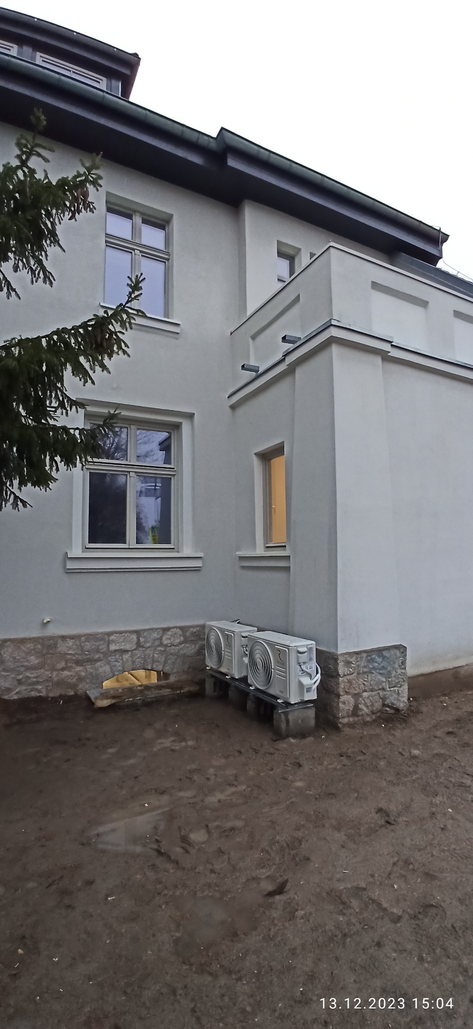 Dwa zewnętrzne agregaty klimatyzacji zamontowane na betonowych podstawach przy ścianie budynku z widocznymi oknami i fragmentem drzewa iglastego.