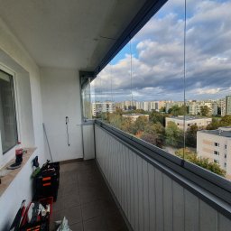 Junior Construction - Zabudowany balkon z widokiem na osiedle, skrzynki z narzędziami i butelka coli przy oknie, szklane panele balkonowe.