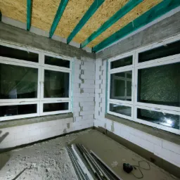 Dwa białe okna PCV zamontowane w surowym wnętrzu z białych bloczków, widoczne elementy konstrukcji dachu z drewnianych belek pomalowanych na zielono, pod oknami leżą profile okienne i narzędzia.