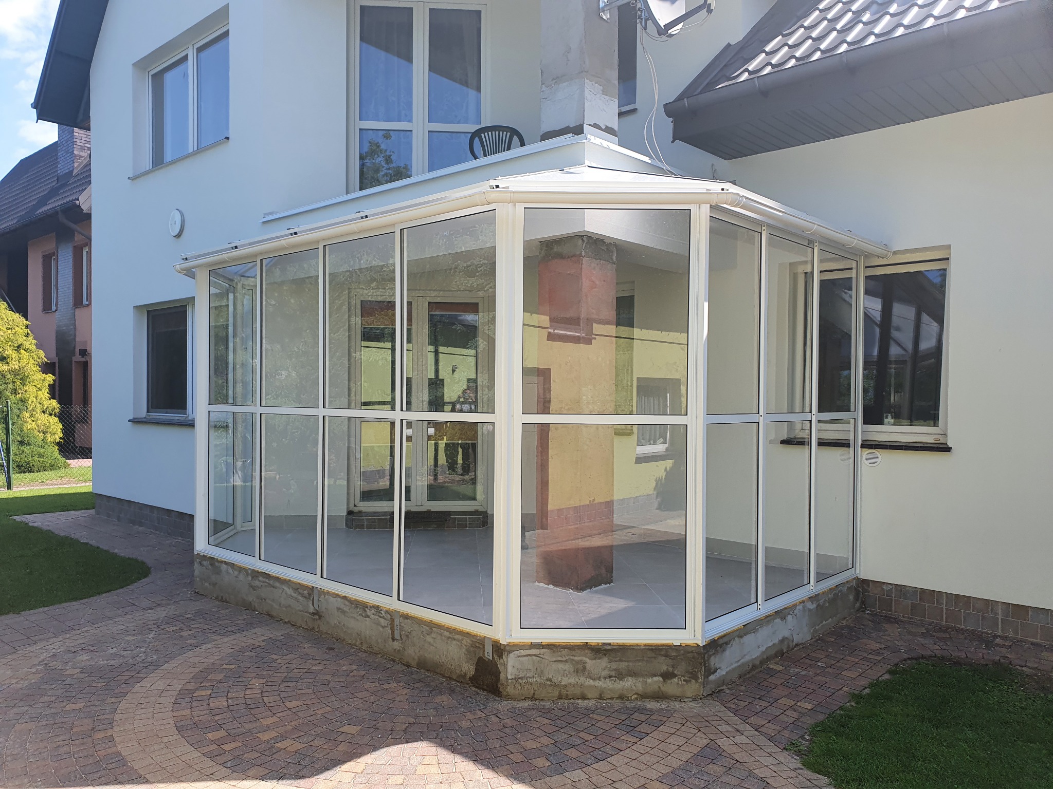 Nowoczesna, przeszklona oranżeria z białymi ramami, przylegająca do białego domu, z widocznym kominem i krzesłem ogrodowym na dachu, otoczona brukowanym tarasem.