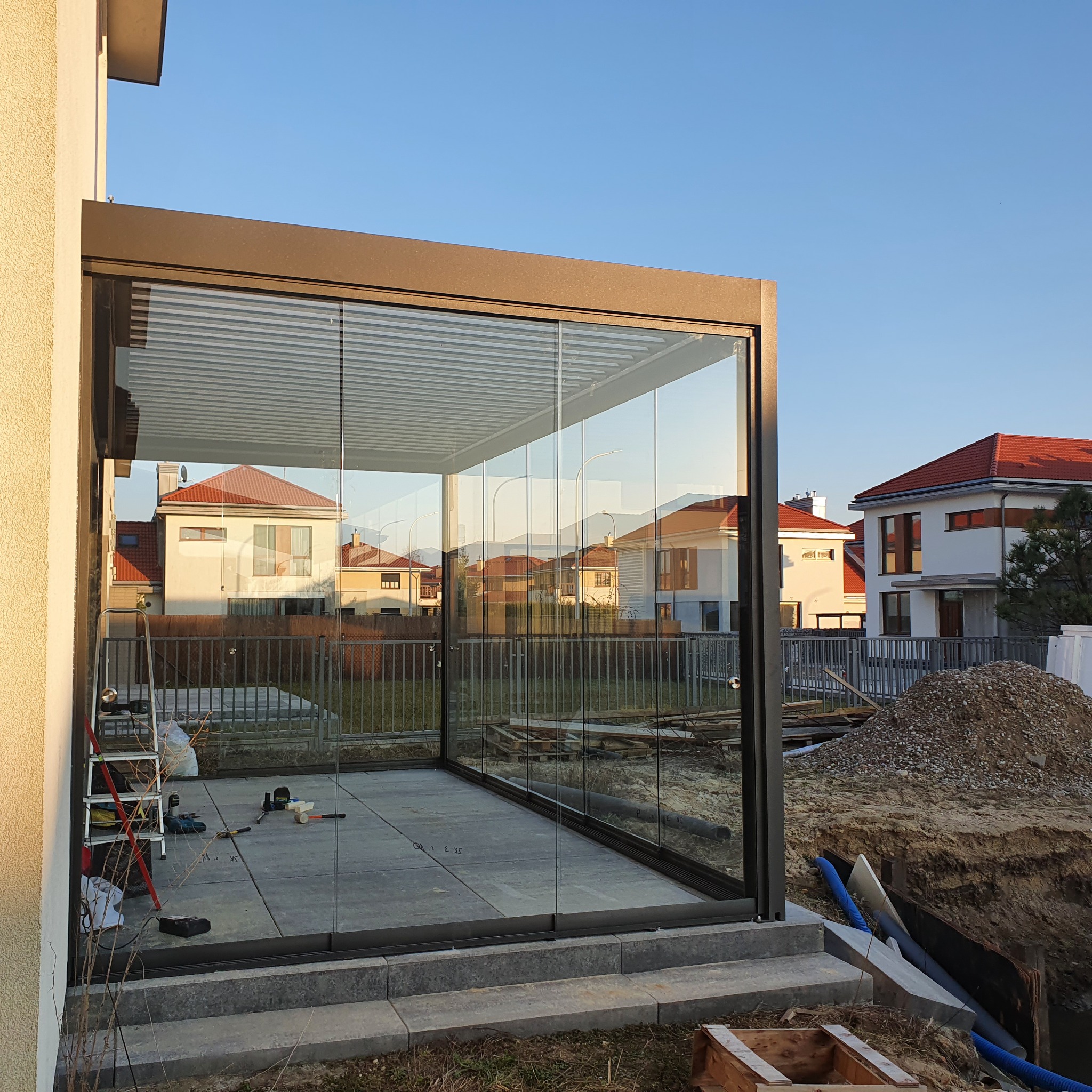 Szklana zabudowa tarasu z przesuwnymi panelami w nowoczesnym domu, widoczne narzędzia i drabina sugerują trwający montaż.