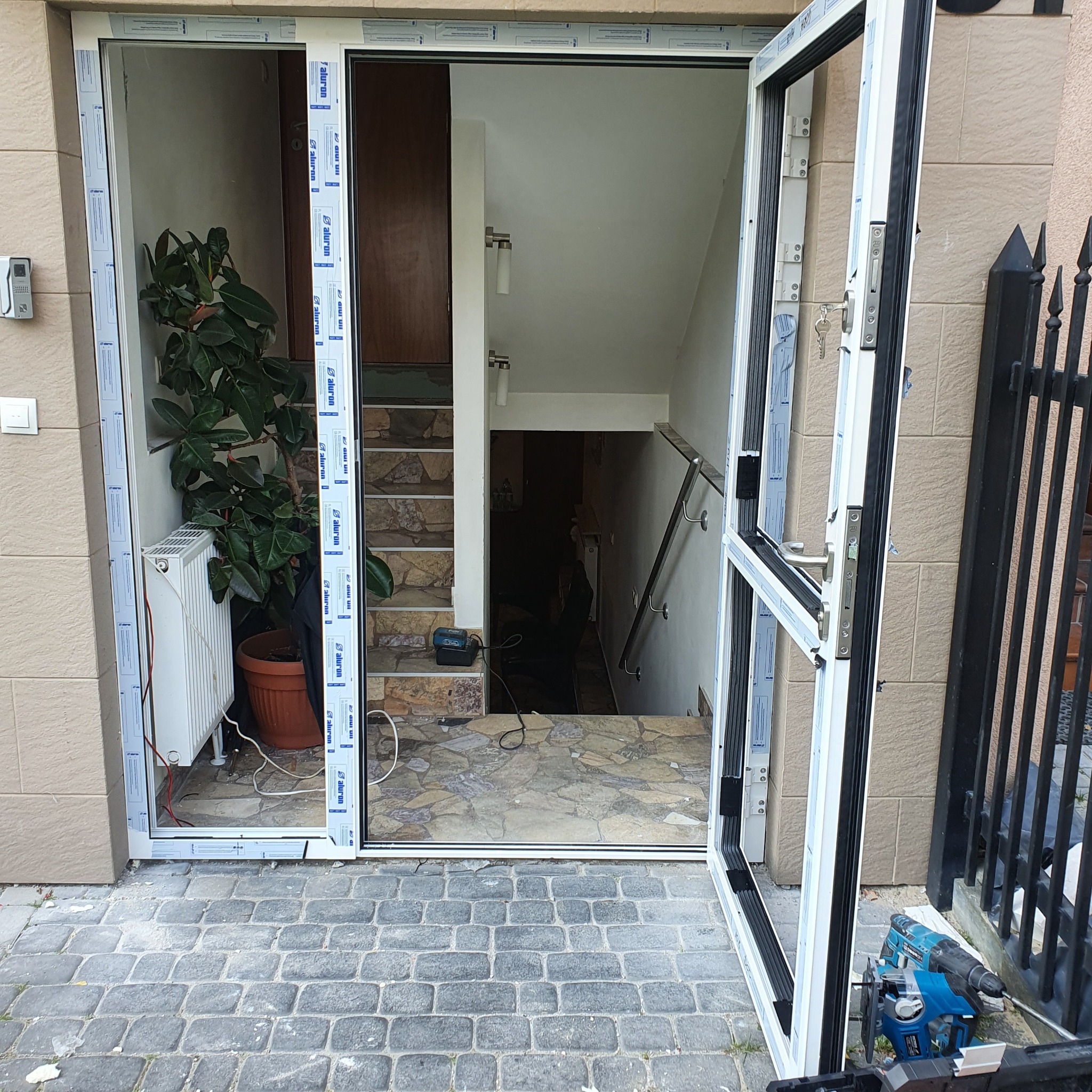 Otwarty, biały, aluminiowy profil okienny z widocznym wnętrzem budynku: schody, poręcz, roślina doniczkowa i narzędzia budowlane, sugerujący etap montażu.