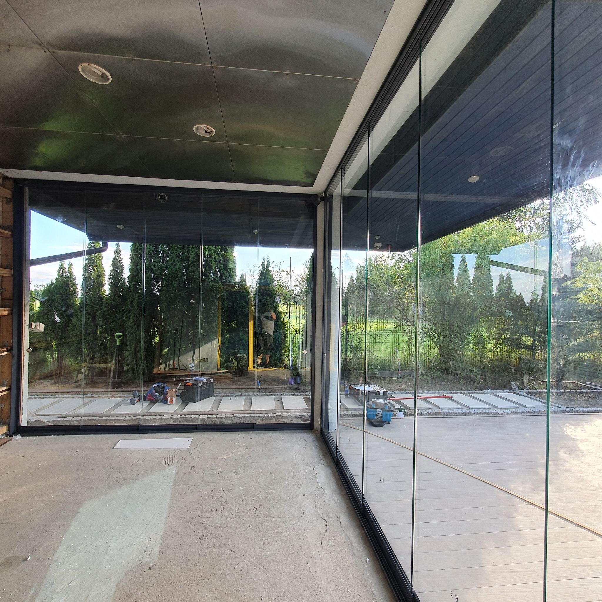 Przesuwne szklane ściany tarasu w trakcie montażu, widoczne narzędzia i materiały budowlane, na zewnątrz zieleń ogrodu.