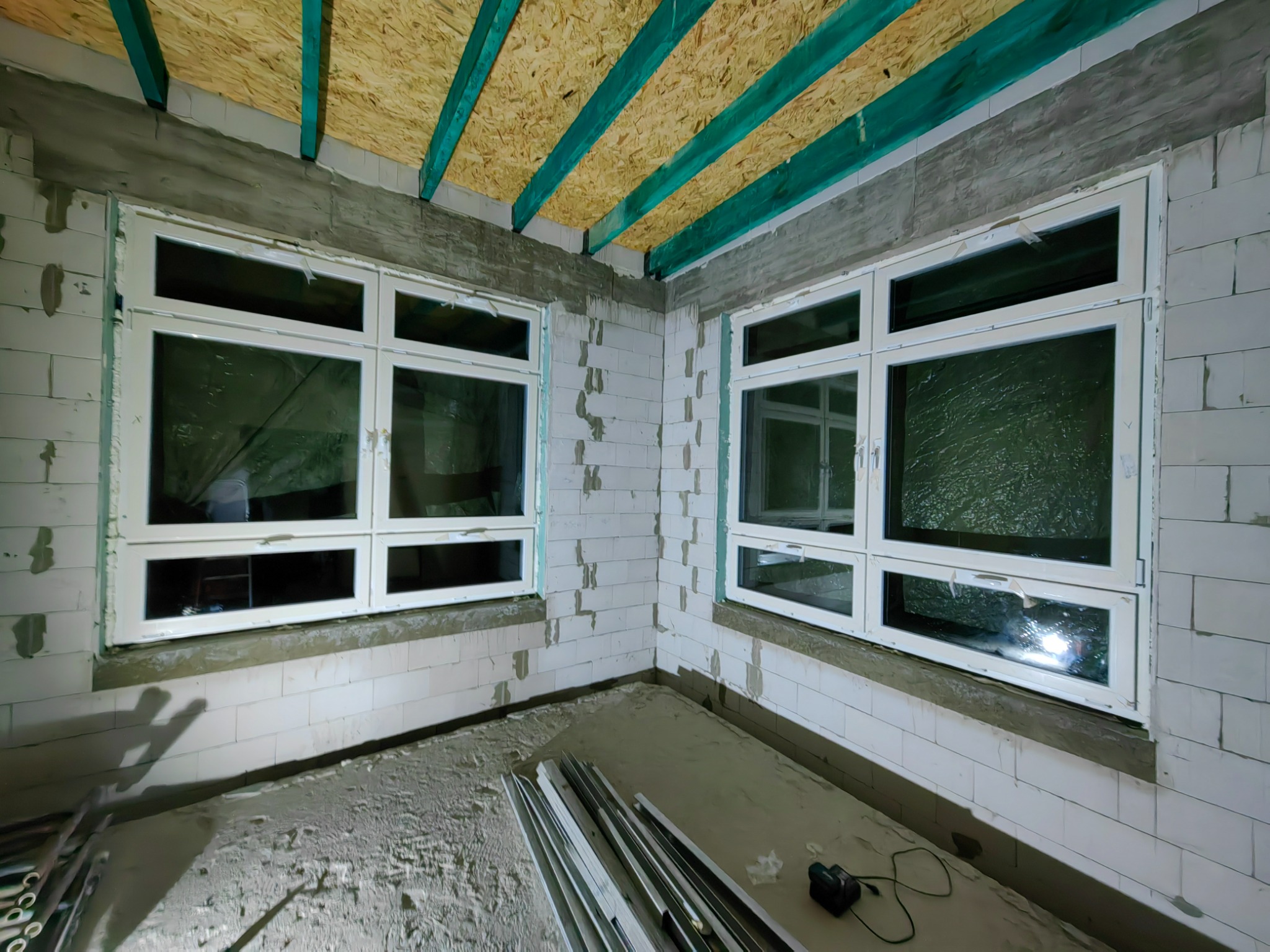 Dwa białe okna PCV zamontowane w surowym wnętrzu z białych bloczków, widoczne elementy konstrukcji dachu z drewnianych belek pomalowanych na zielono, pod oknami leżą profile okienne i narzędzia.