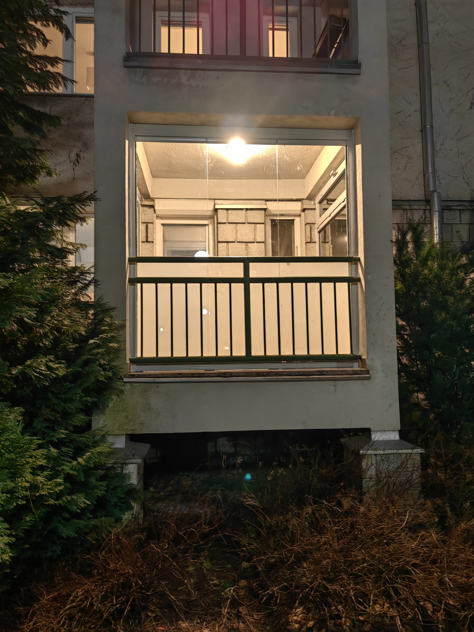 Zabudowany balkon z aluminiowymi ramami i poręczą, widoczny z zewnątrz w wieczornym oświetleniu, otoczony zielenią.