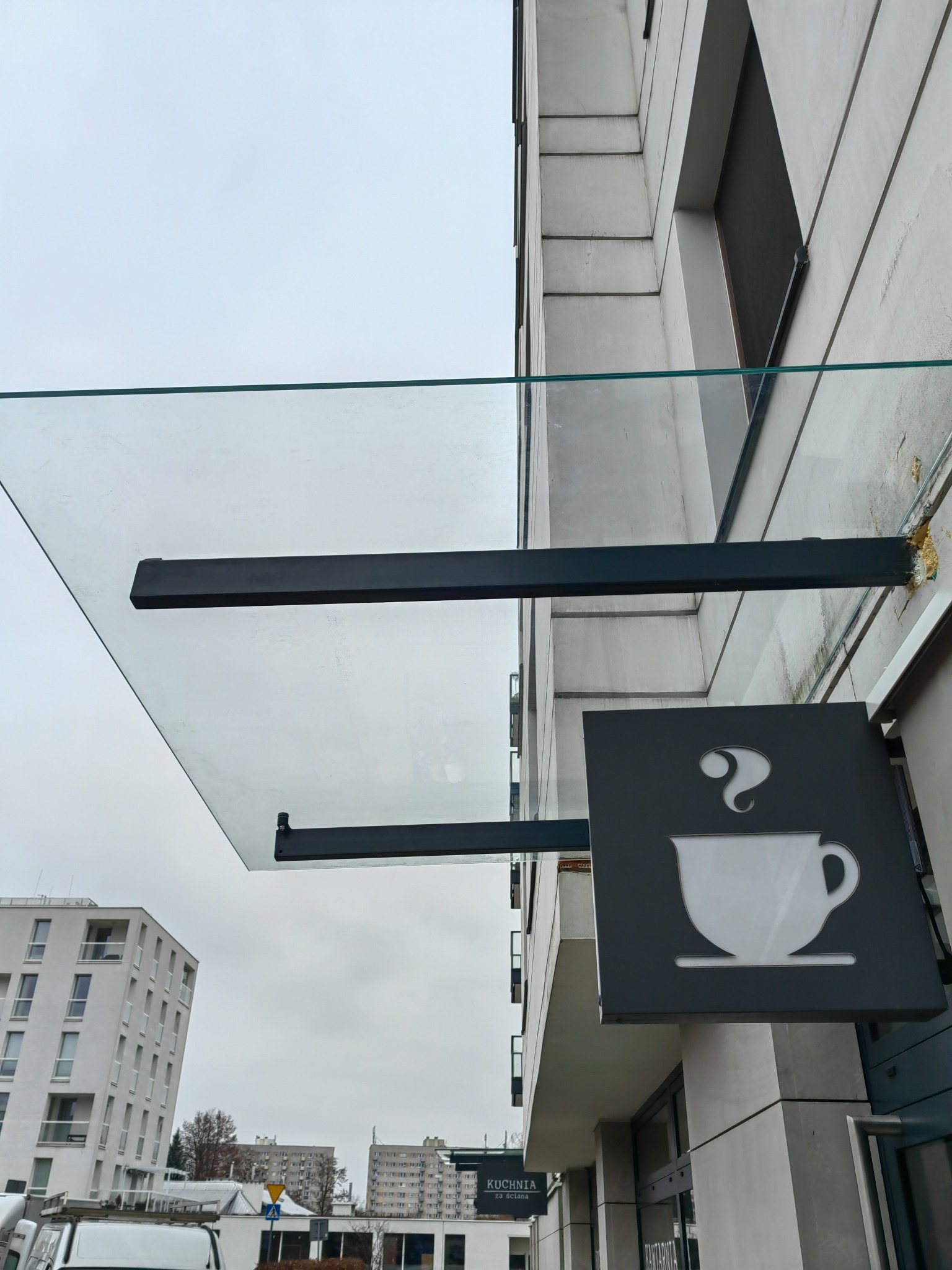 Szklany daszek nad wejściem do kawiarni z logo filiżanki kawy na szyldzie, widoczne budynki w tle.