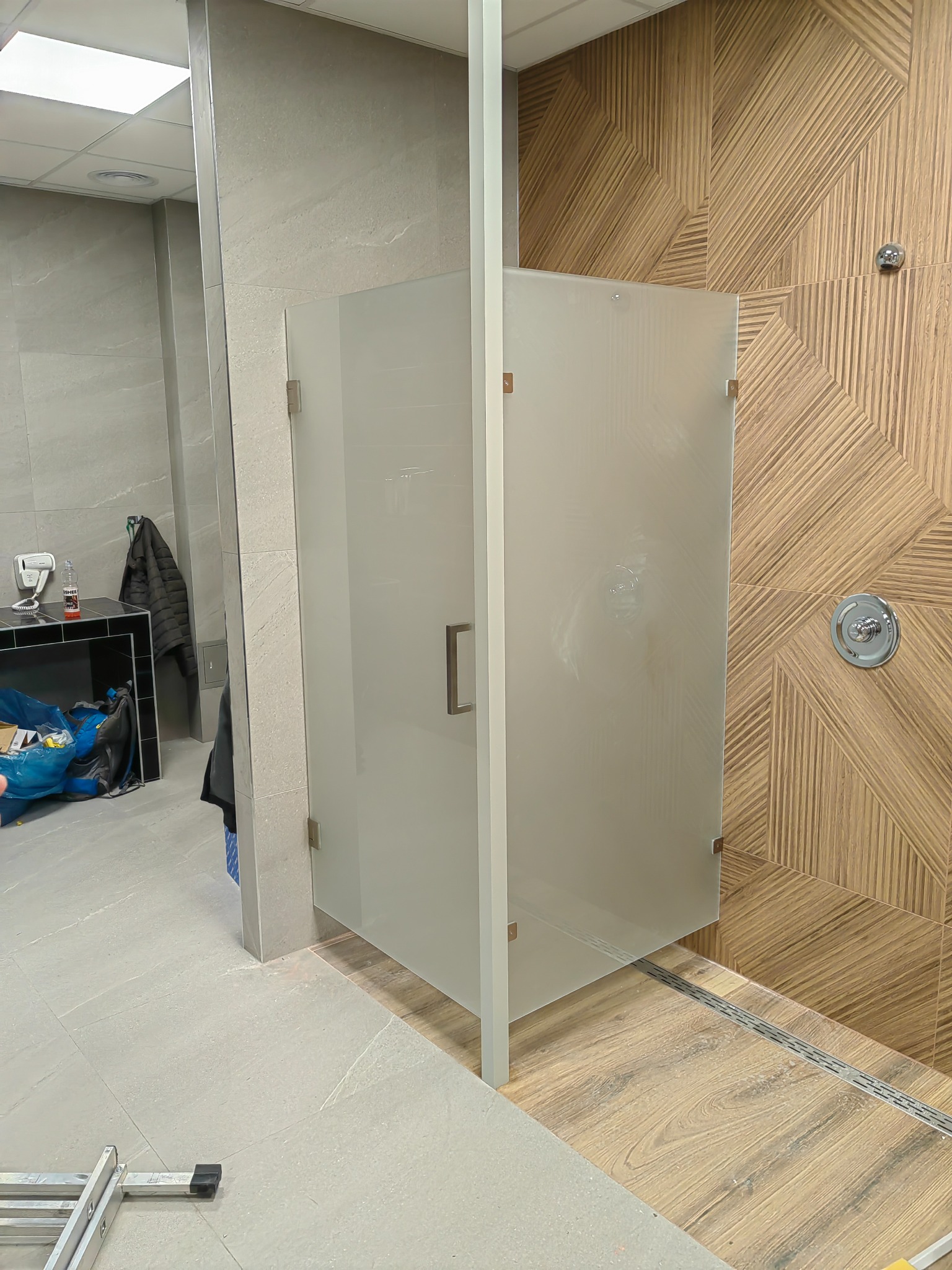 Szklana kabina prysznicowa z matowymi drzwiami zamontowana w łazience z szarą i drewnopodobną okładziną ścienną, widoczny odpływ liniowy w podłodze i drabina w rogu.