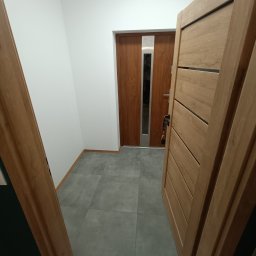 Stefmarc Spółka z Ograniczoną Odpowiedzialnością - Wnętrze przedpokoju z szarą, kwadratową podłogą z płytek, białe ściany i dwoje drewnianych drzwi: otwarte drzwi po prawej i zamknięte, brązowe drzwi wejściowe z pionowym, metalowym panelem po lewej.