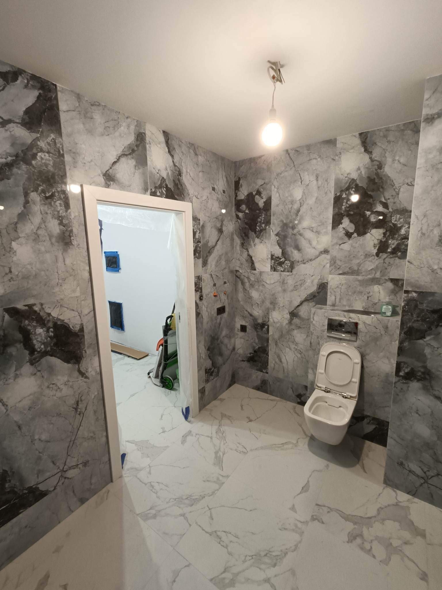 Wykończona łazienka z marmurowymi płytkami w odcieniach szarości i bieli, wisząca toaleta z otwartą klapą, widok na sąsiednie pomieszczenie przez zabezpieczone folią drzwi, gdzie widoczny sprzęt...