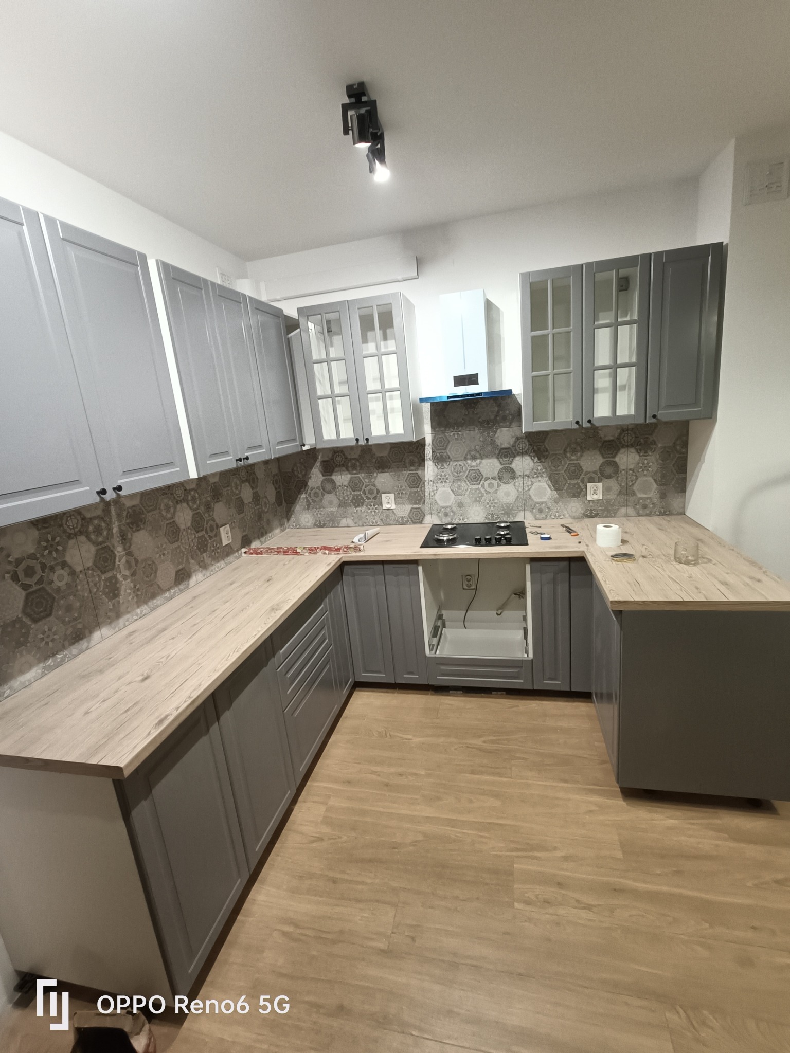 Szara kuchnia w trakcie montażu: szafki wiszące z przeszklonymi frontami, heksagonalne płytki nad blatem, płyta gazowa i brakujące drzwi szafki pod zlewem.