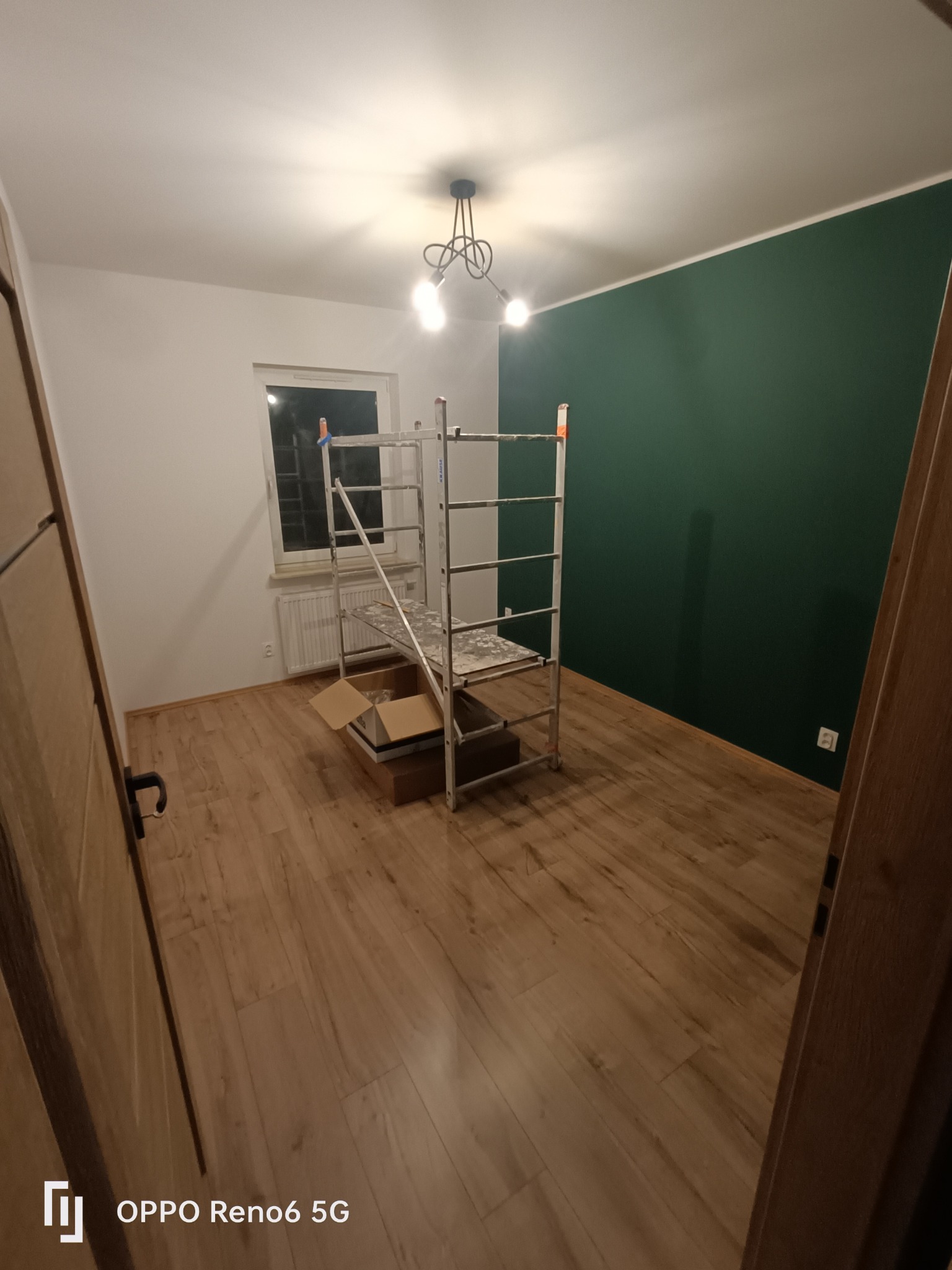 Świeżo pomalowane wnętrze w Warszawie: jedna ściana w kolorze butelkowej zieleni, pozostałe białe, podłoga wyłożona panelami, widoczne rusztowanie i kartony, sugerujące trwający remont.