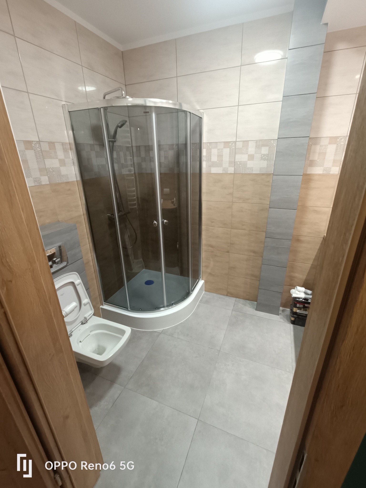 Wykończona łazienka z narożną kabiną prysznicową z ciemnego szkła, wiszącą toaletą z otwartą deską, szarymi płytkami podłogowymi i wielokolorowymi płytkami ściennymi z dekoracyjnymi wstawkami.