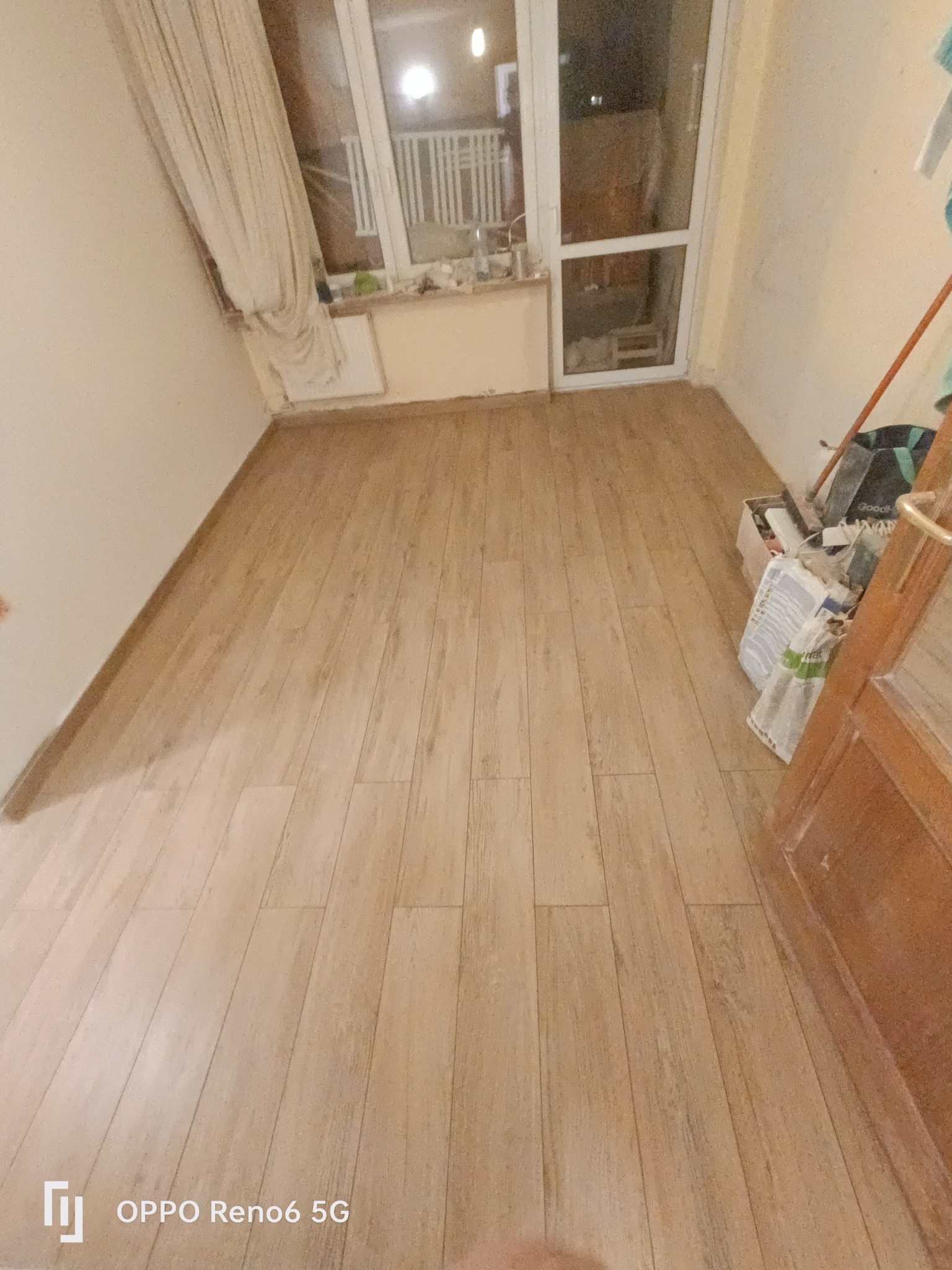 Ułożone panele podłogowe w jasnym odcieniu drewna, widoczne przy oknie z zasłoną i ścianie z narzędziami w trakcie prac wykończeniowych.