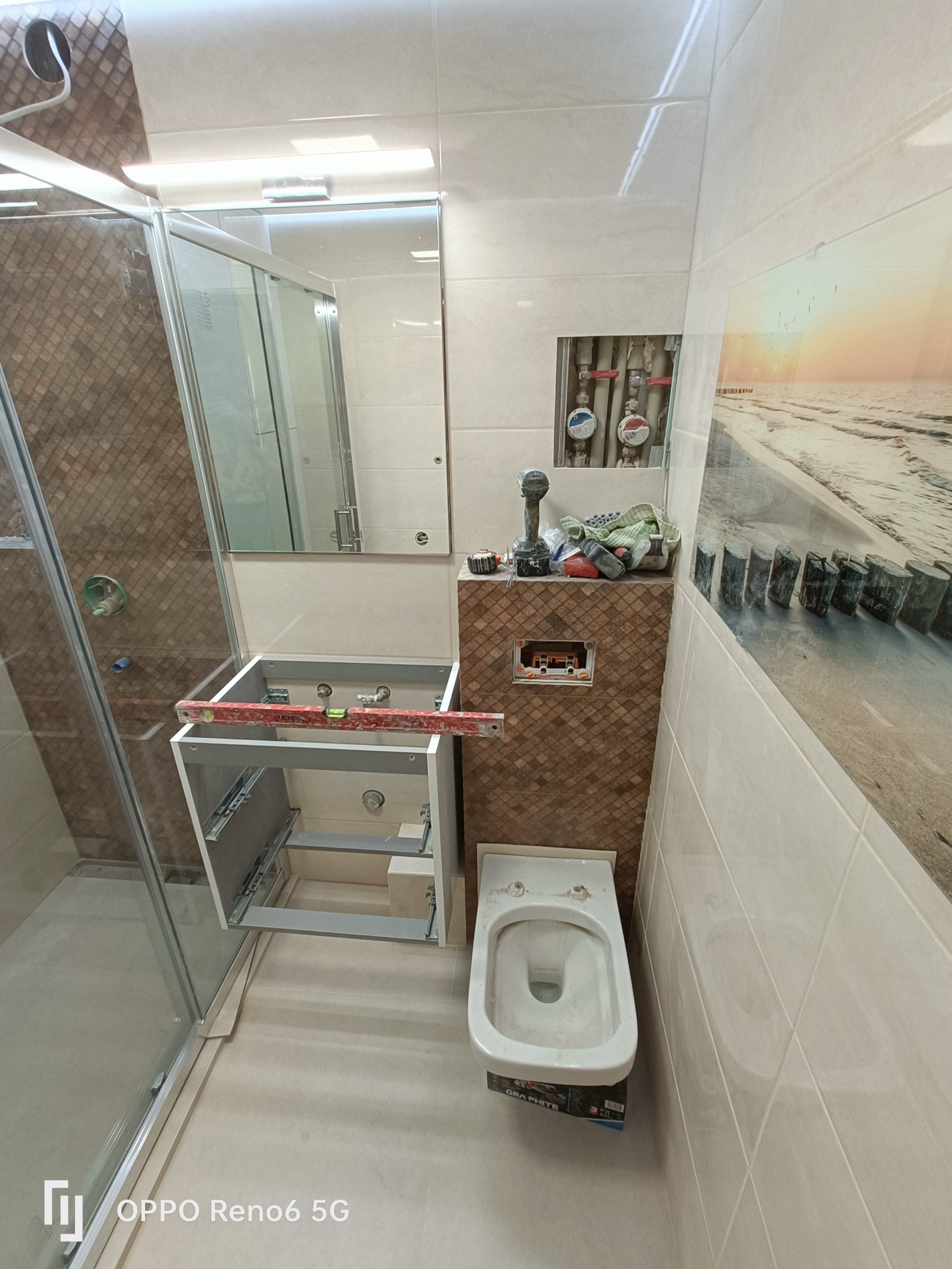 Montaż szafki pod umywalkę w łazience z widocznym poziomowaniem, obok toaleta i kabina prysznicowa, na ścianie płytki imitujące drewno i obraz z motywem morskim.