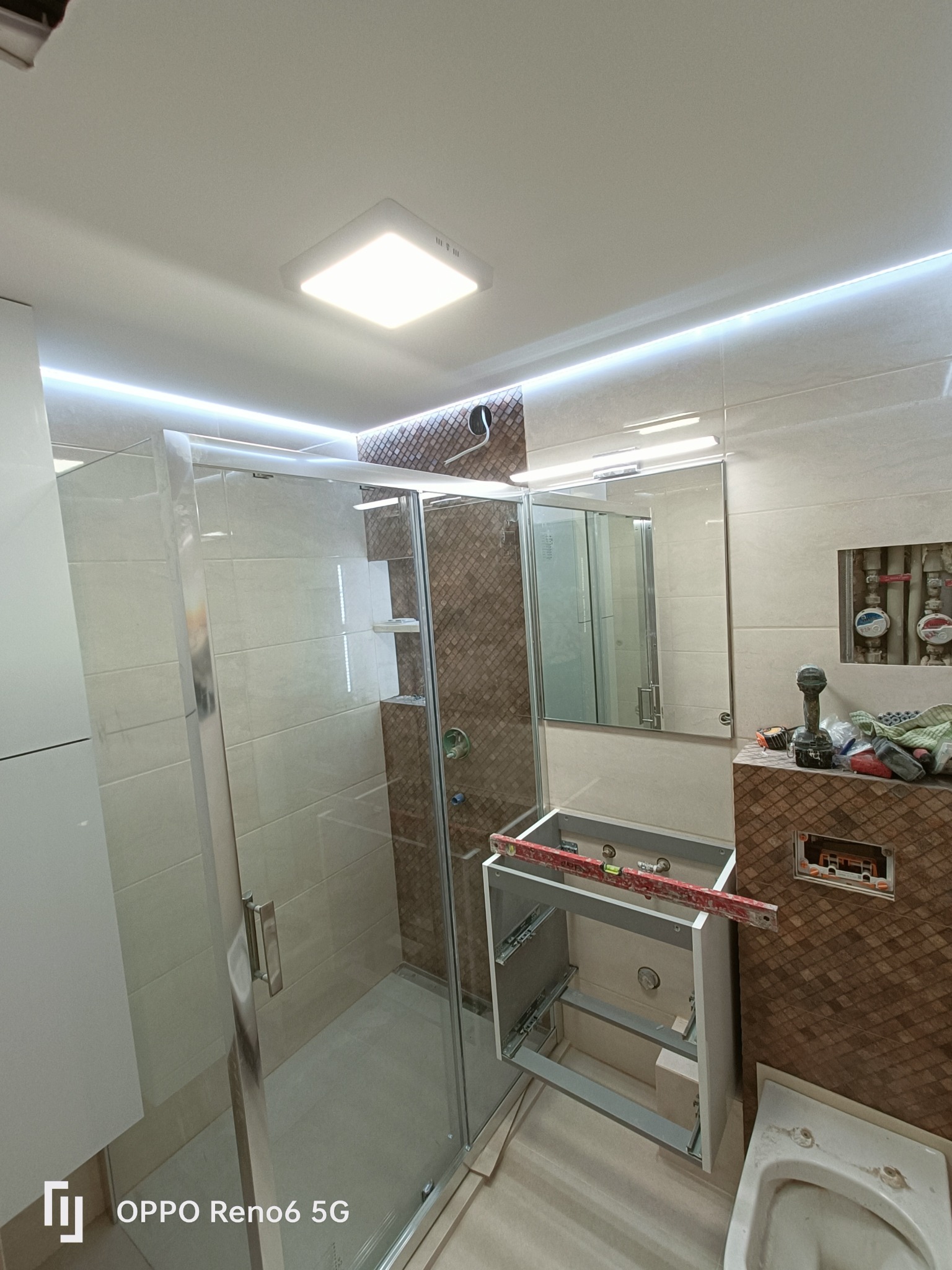 Wnętrze łazienki w trakcie prac: kabina prysznicowa z półkami, częściowo zamontowana szafka pod umywalkę z poziomicą, płytki imitujące drewno na ścianie, widoczne rury i narzędzia.