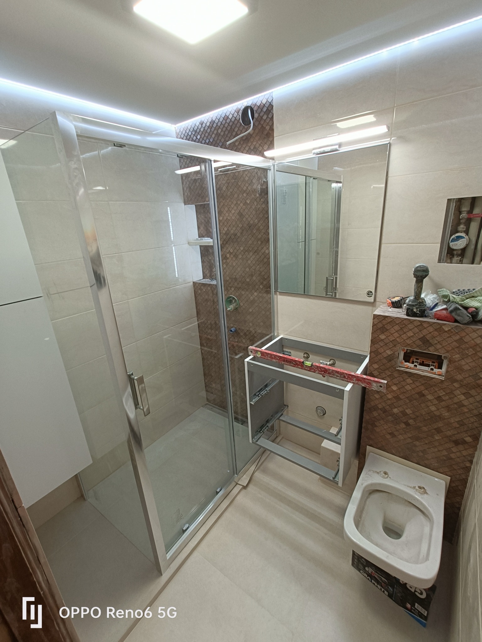 Wnętrze łazienki w trakcie prac: kabina prysznicowa ze srebrnymi profilami, niedokończona szafka pod umywalkę z poziomicą, przygotowana do montażu toaleta na kartonie, oraz narzędzia na tle mozaiki...