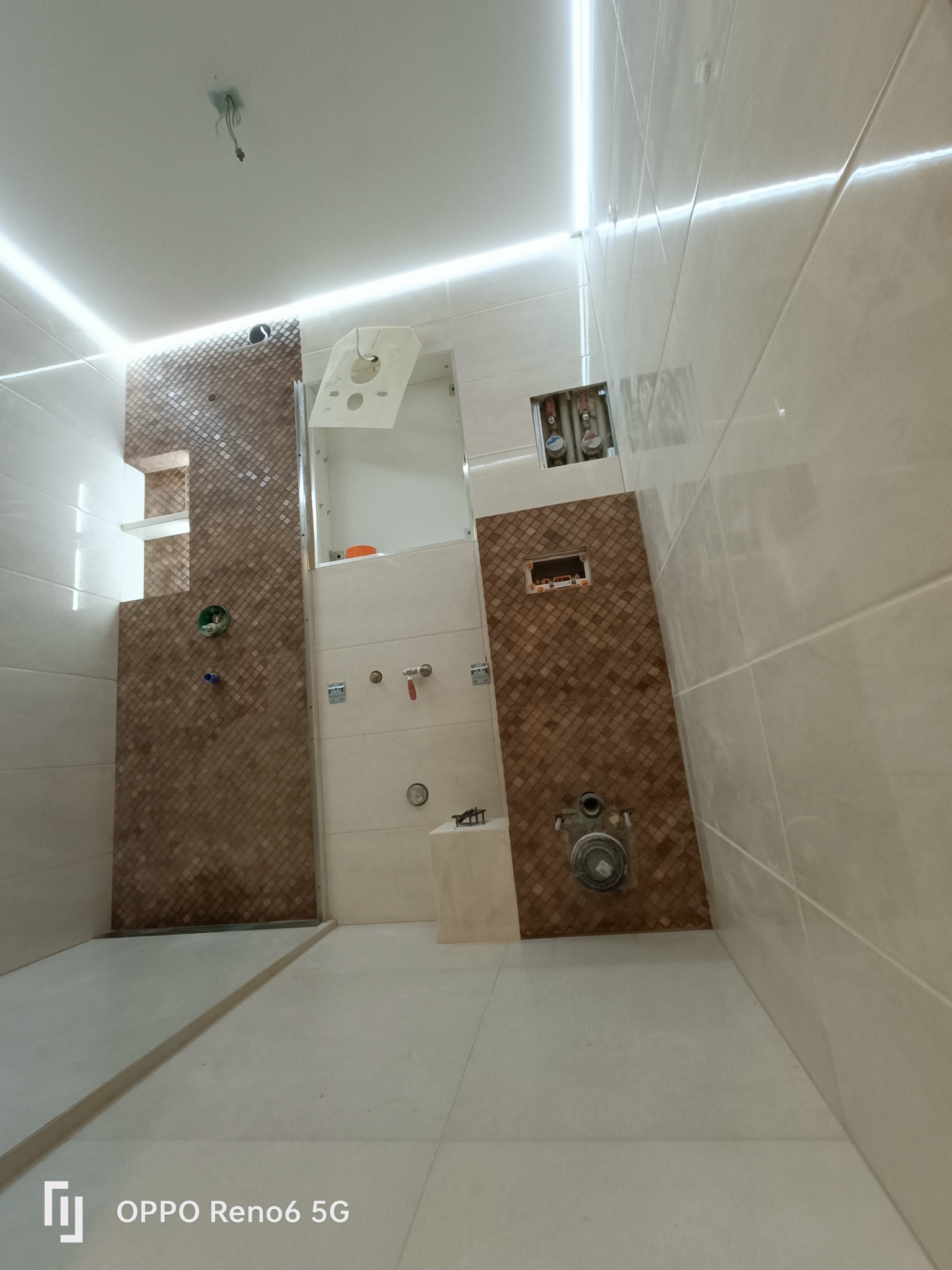 Wnętrze łazienki w trakcie prac: częściowo ułożone beżowe płytki na ścianach i podłodze, fragment ściany z dekoracyjnymi brązowymi płytkami mozaikowymi, widoczne instalacje wodno-kanalizacyjne...